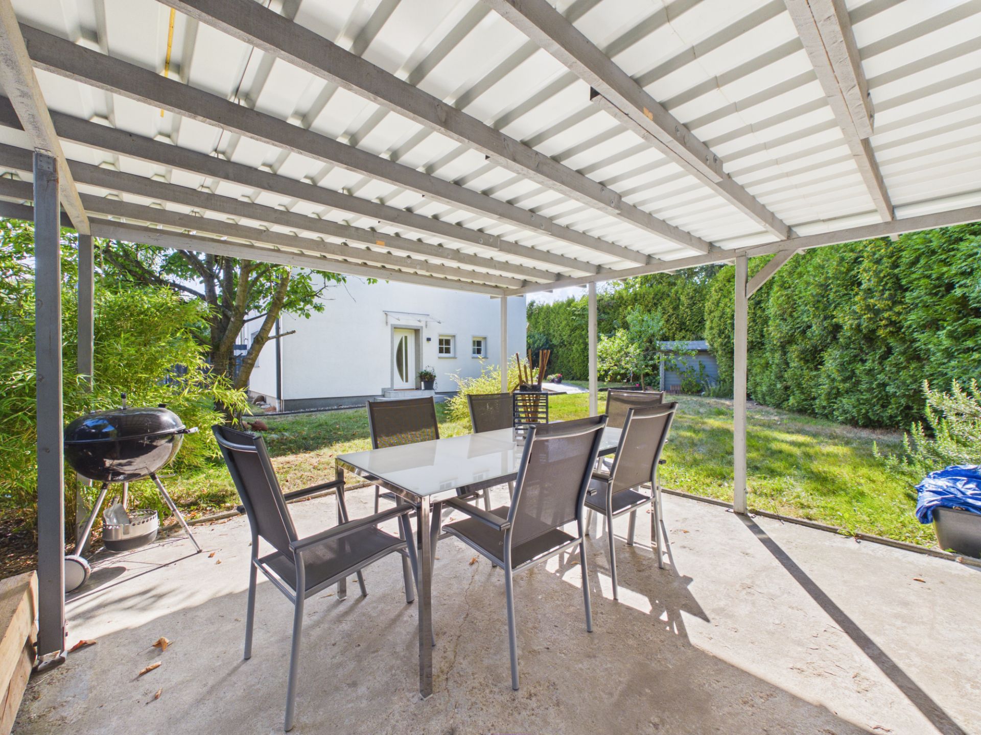 Überdachte Terrasse mit Gartentisch, sechs Stühlen und Grill im Garten eines Wohnhauses.