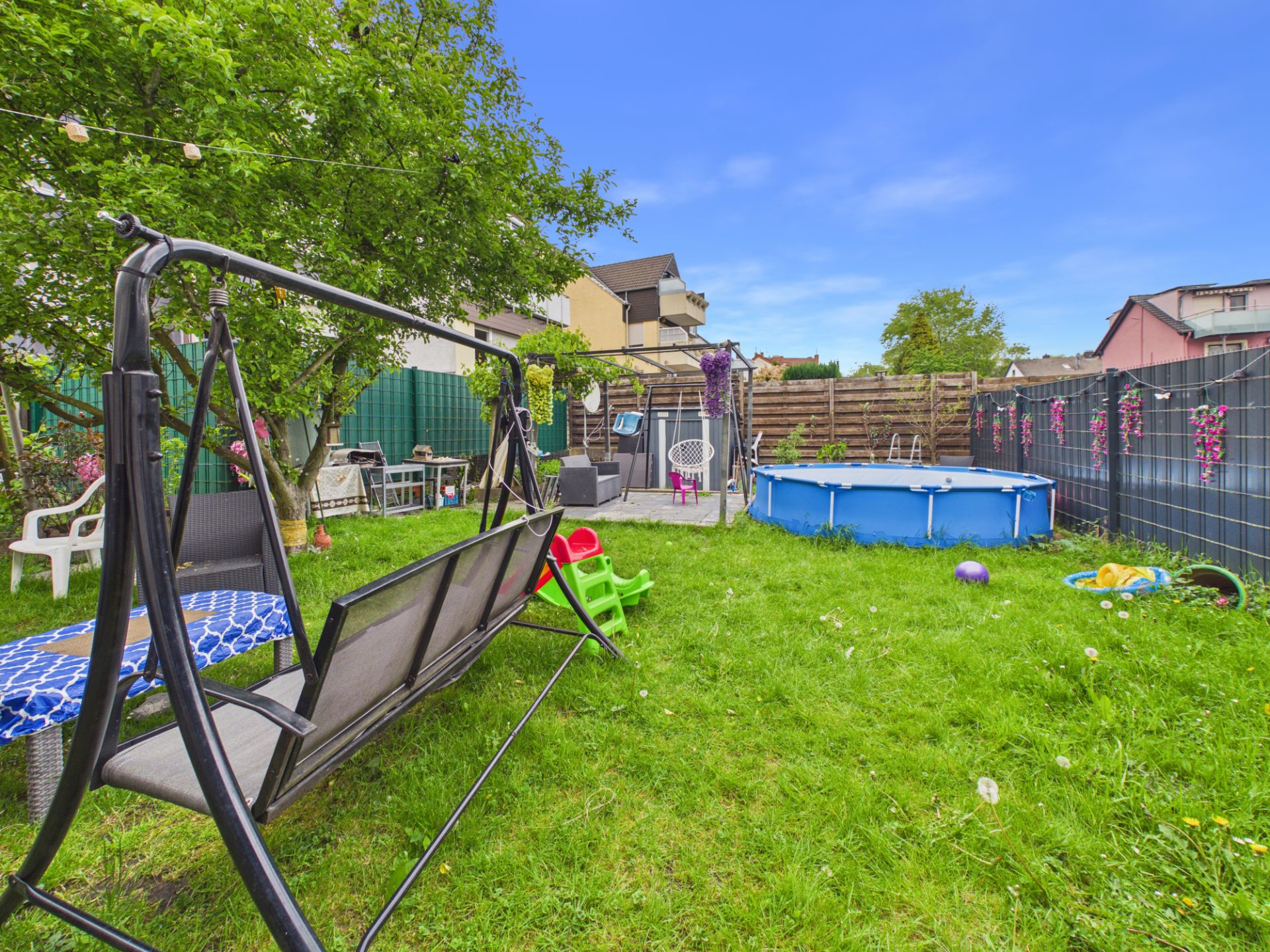Garten mit Schaukelbank, aufgestelltem Pool, Gartenmöbeln und Spielzeug auf grünem Rasen.