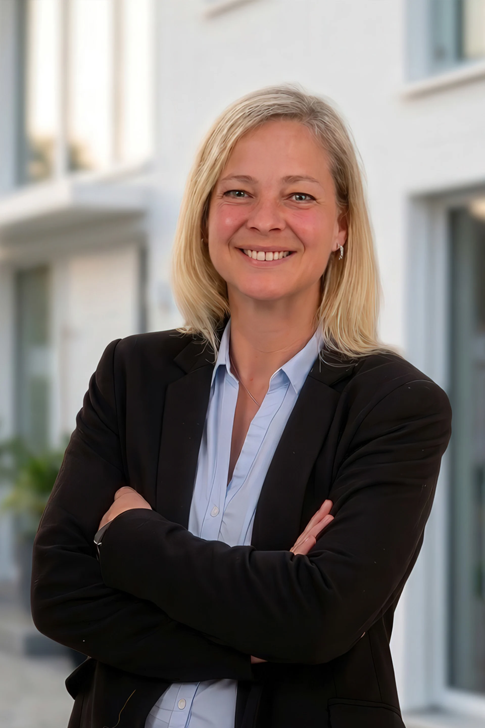 Frau mit blonden Haaren und Anzug steht lächelnd vor einem modernen Wohngebäude.