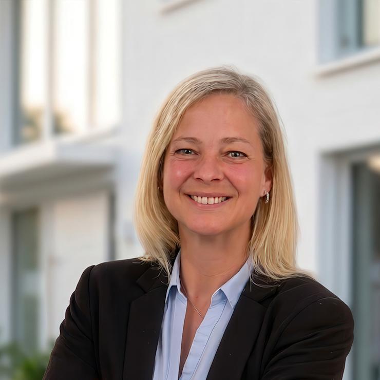 Frau mit blonden Haaren und Anzug steht lächelnd vor einem modernen Wohngebäude.