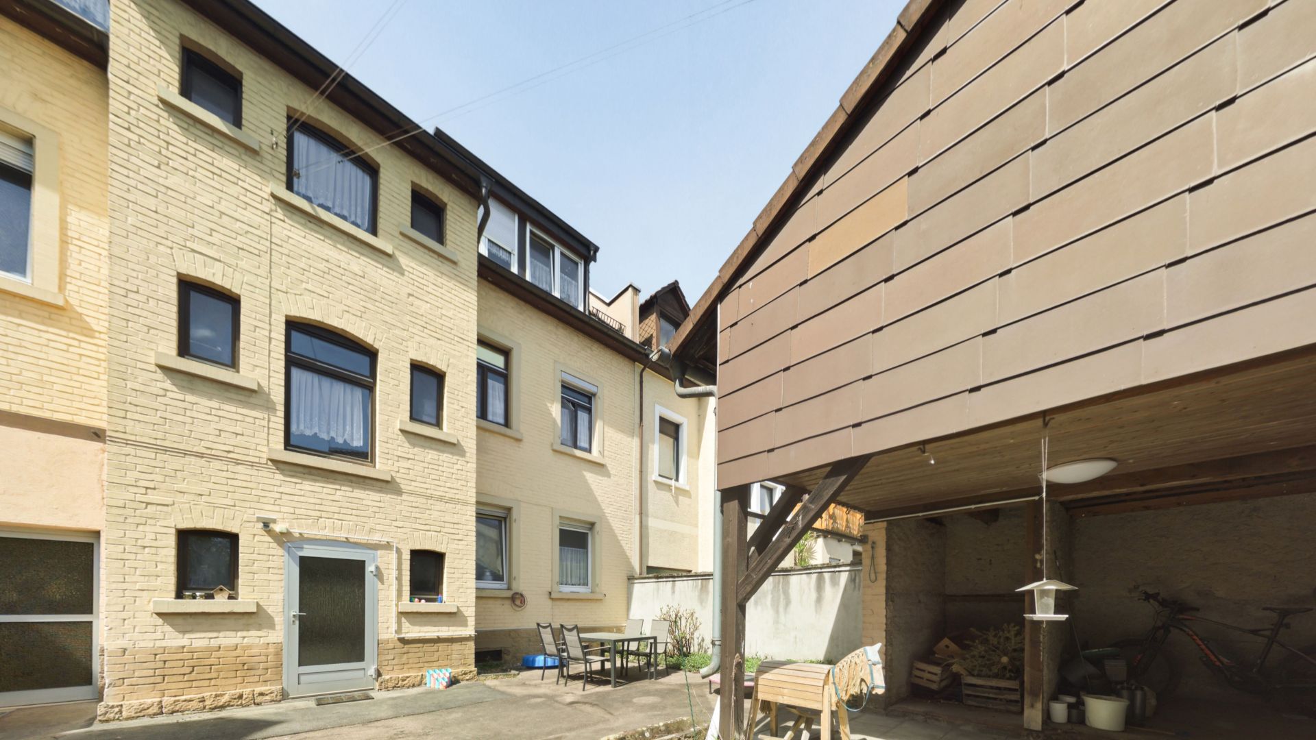 Innenhof mit mehrstöckigem Wohnhaus, überdachtem Holzunterstand und Gartenmöbeln.