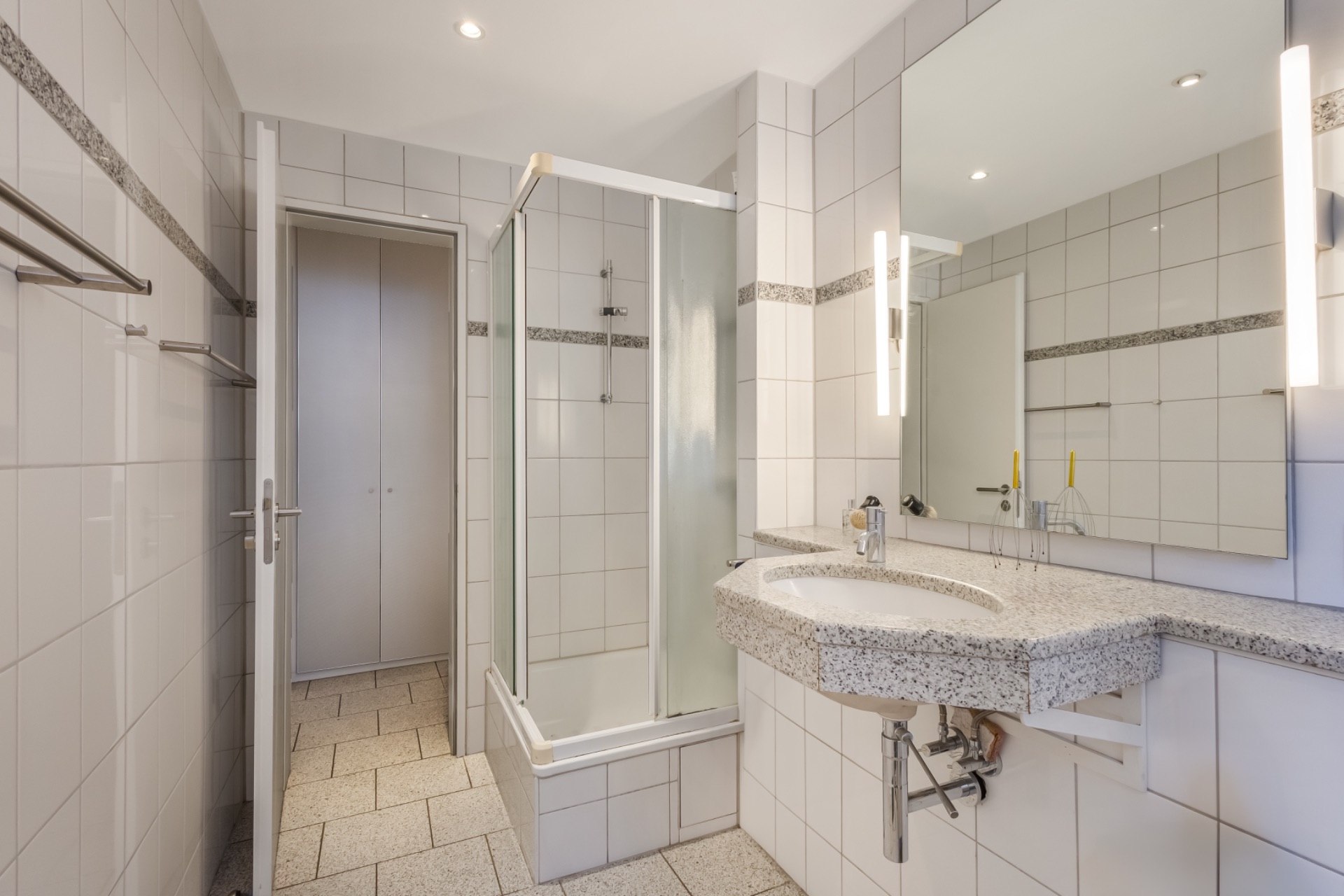 Modernes, weiß gefliestes Bad mit Dusche, Spiegel, Waschbecken und Handtuchhaltern.