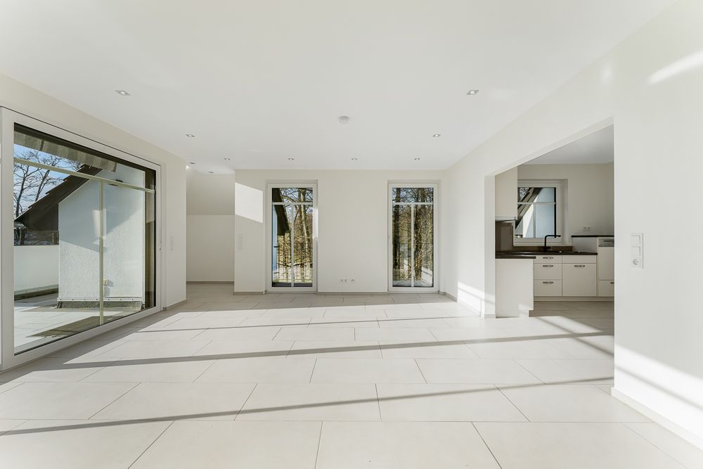 Heller, leerer Wohnraum mit großen Fenstern und Zugang zu Küche und Terrasse.