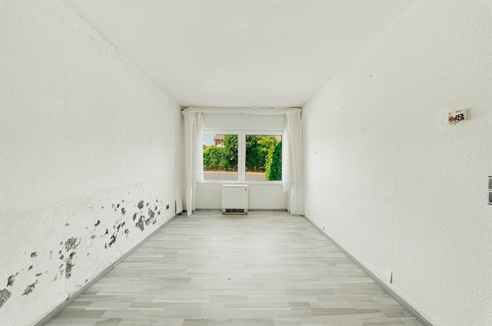 Leerer Raum mit hellem Boden, Fenster, weißen Wänden und sichtbaren Schäden an einer Wand.