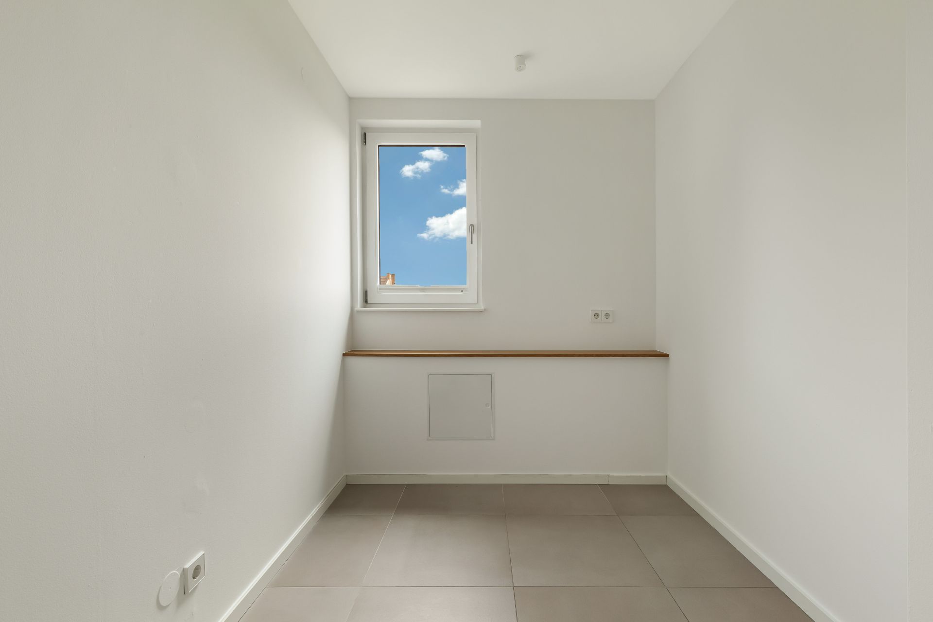 Leerer, heller Raum mit grauen Fliesen und einem Fenster, durch das blauer Himmel zu sehen ist.