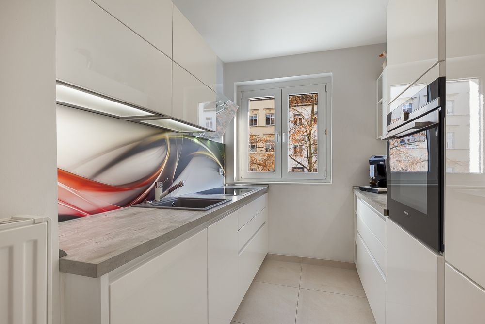 Moderne, helle Einbauküche mit Fenster, Einbauschränken und bunter Glasrückwand.
