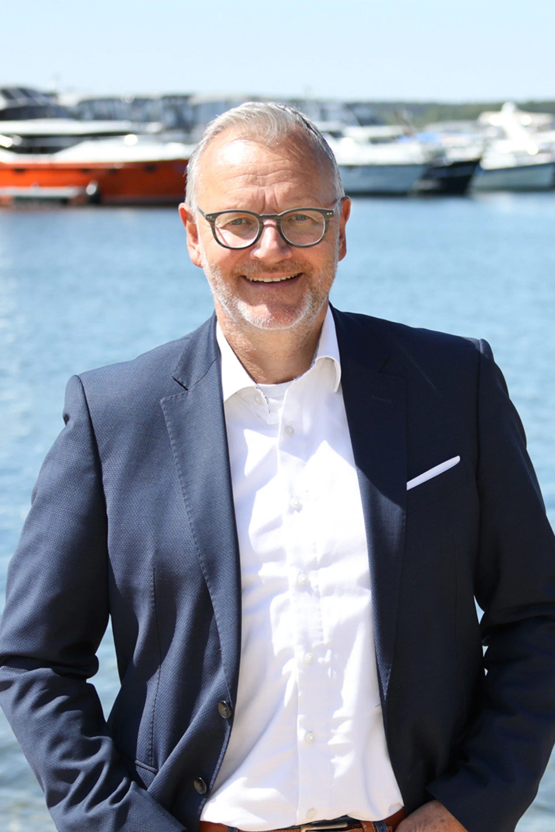 Ein Mann mit Brille im Anzug steht lächelnd vor Wasser mit mehreren Booten im Hintergrund.