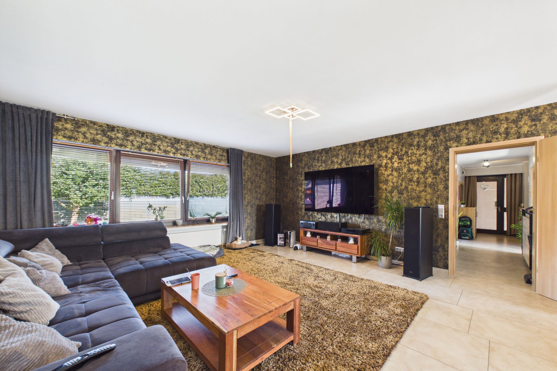 Wohnzimmer mit Ecksofa, Couchtisch, Teppich, großem Fenster und Flachbildfernseher an der Wand.
