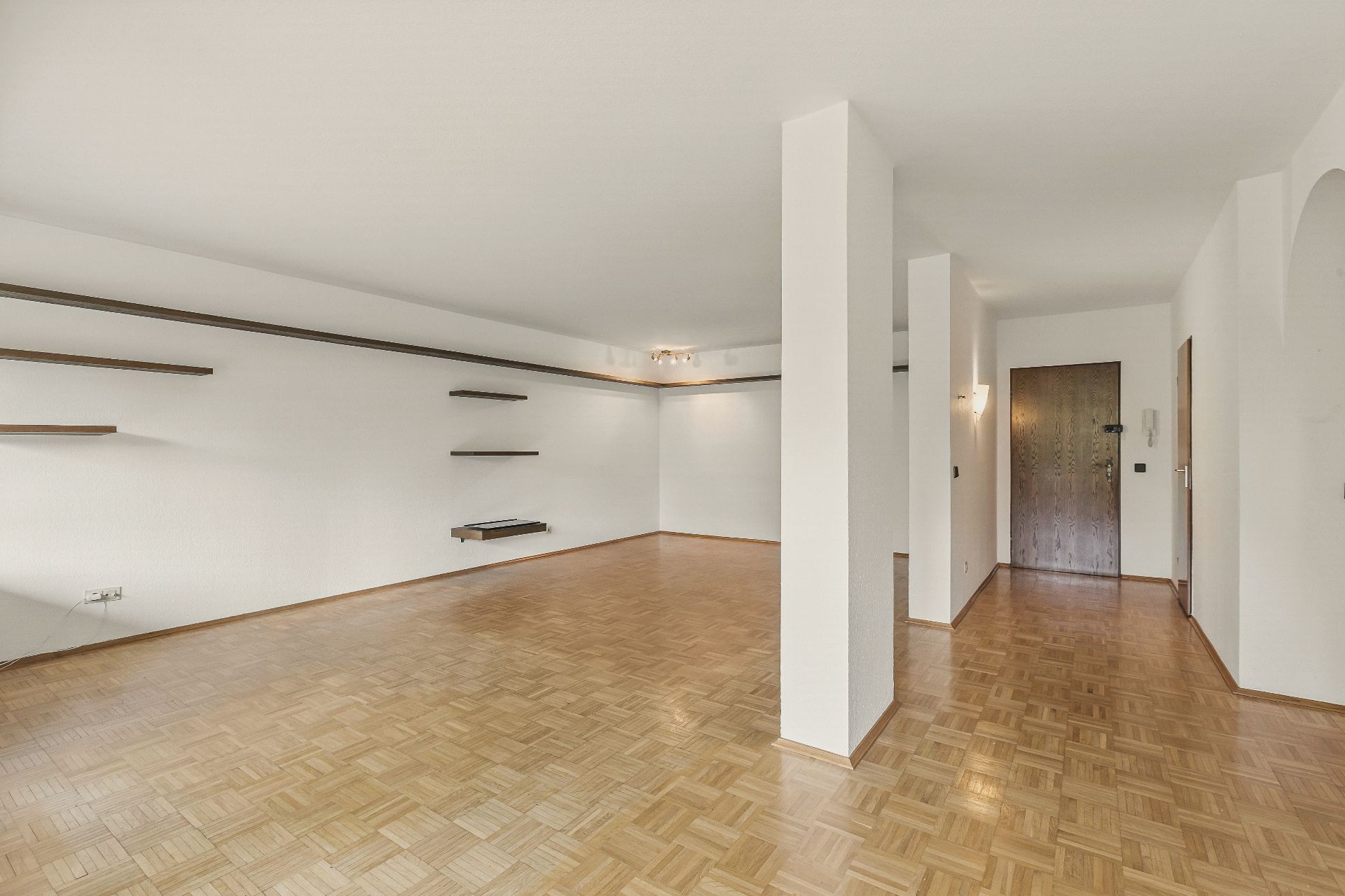 Großes, leeres Wohnzimmer mit Parkettboden, weißen Wänden und mehreren Wandregalen.