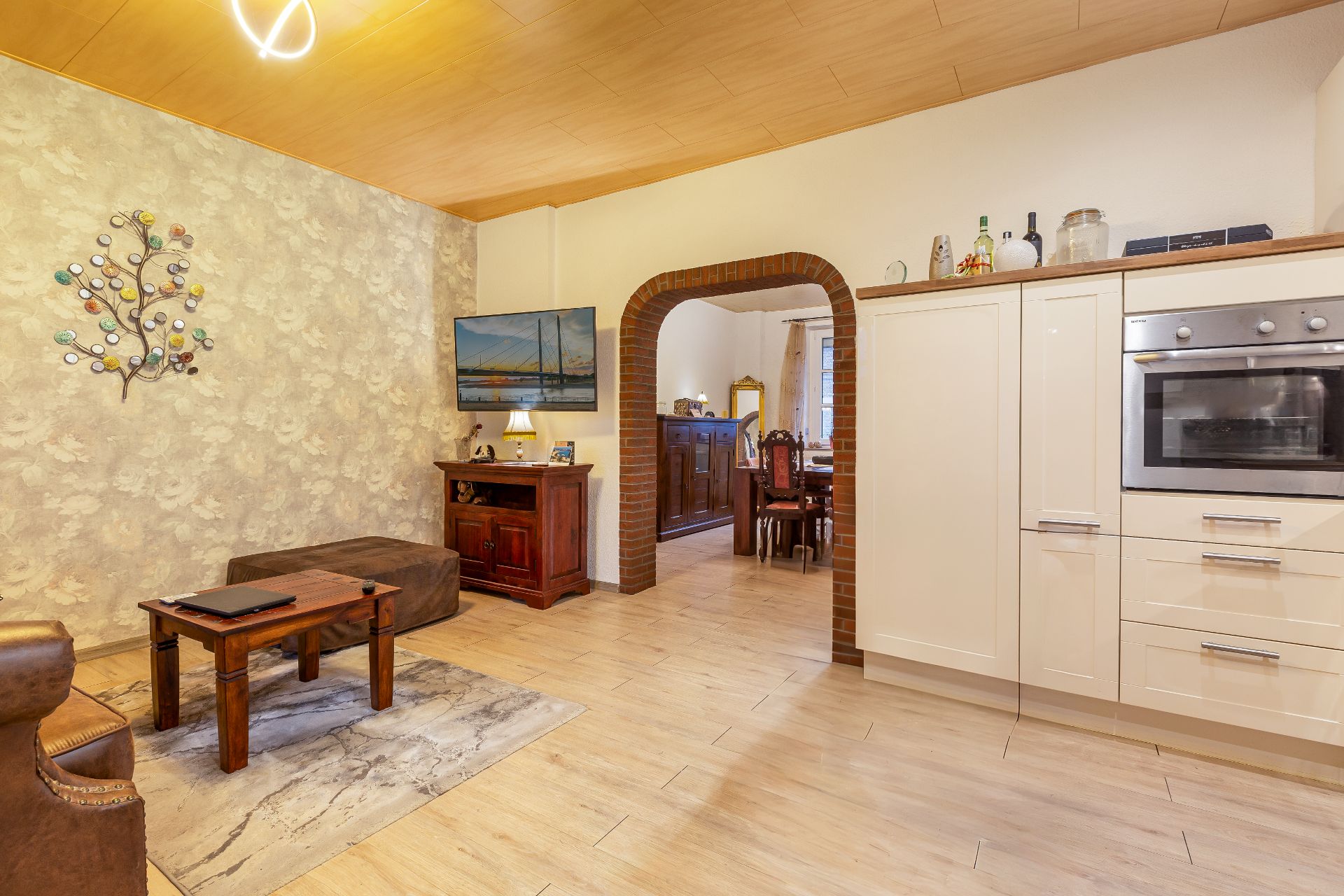 Gemütliches Wohnzimmer mit Holzmöbeln, Wanddeko, Fernseher und Blick ins Esszimmer durch einen Rundbogen.