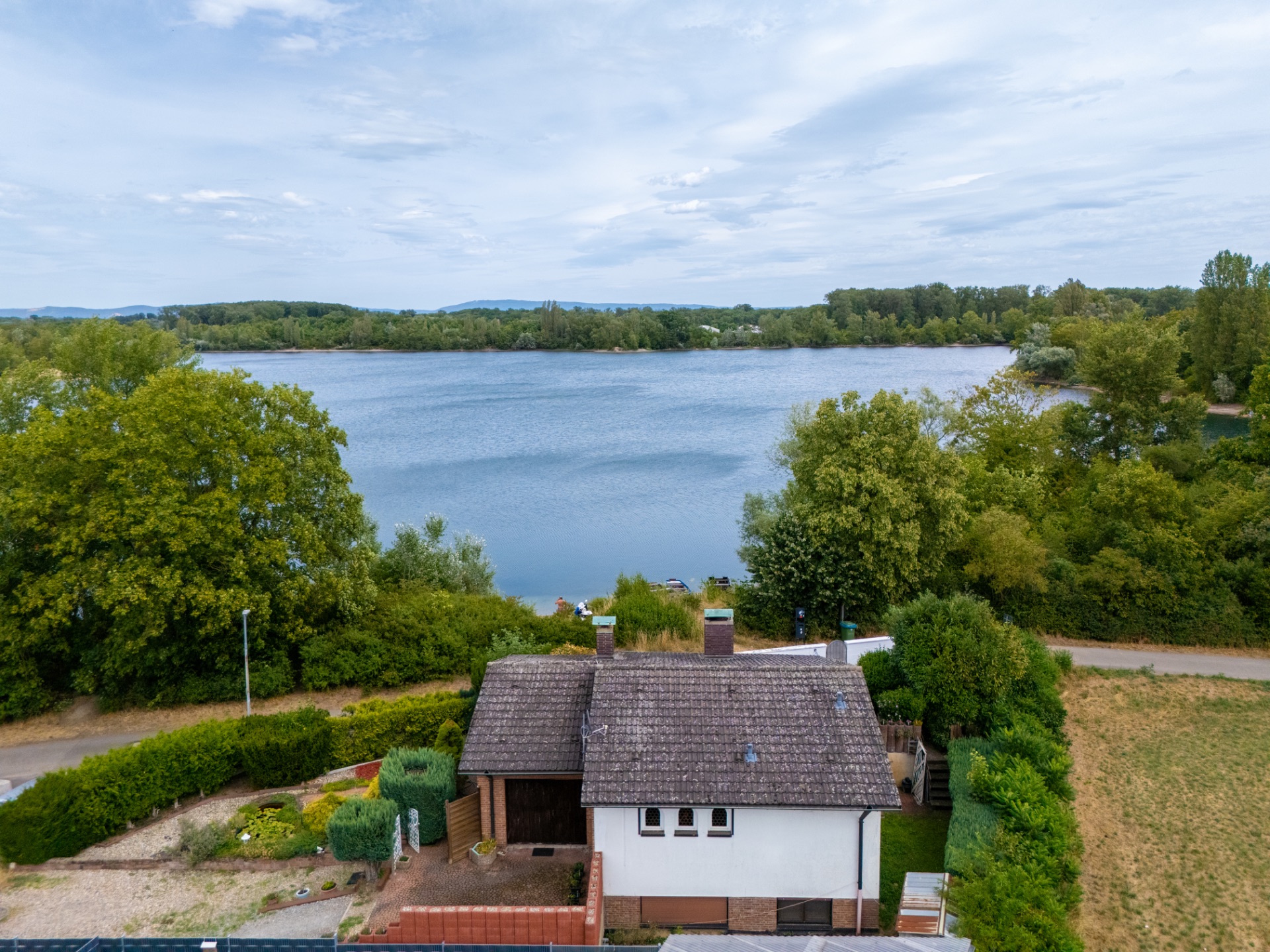 Haus mit Garten am Seeufer, umgeben von Bäumen, mit Blick auf das Wasser und den Himmel.