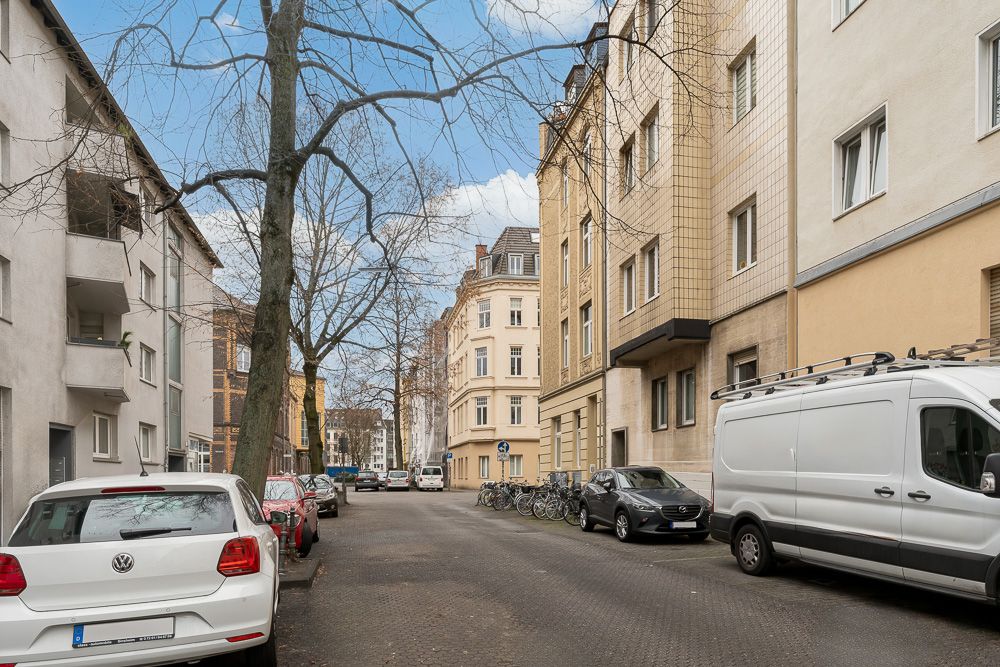 Wohnstraße mit geparkten Autos, Fahrrädern und mehrstöckigen Wohnhäusern an beiden Seiten.
