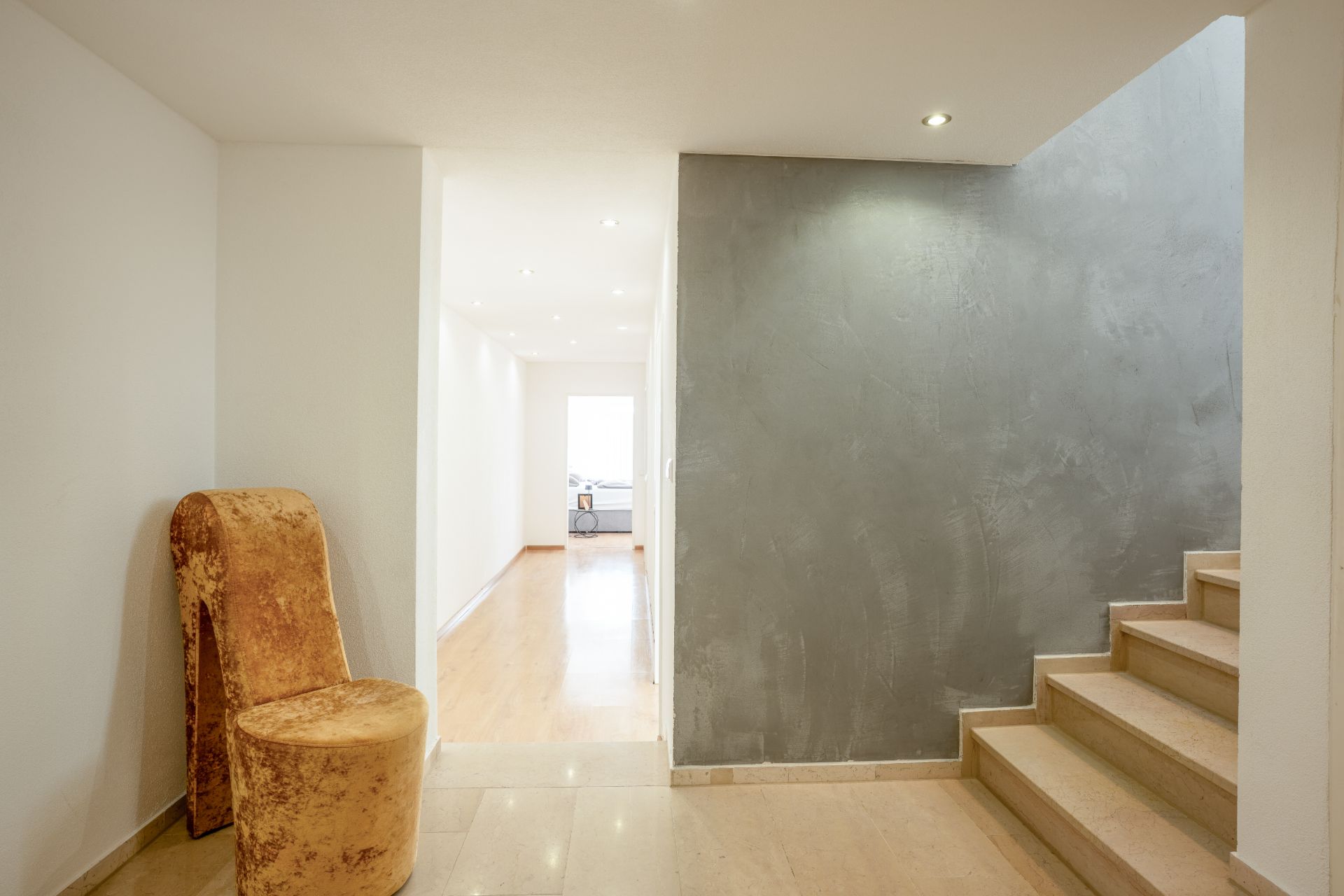 Heller Flur mit Treppe, grauer Wand und einem goldfarbenen Stuhl.