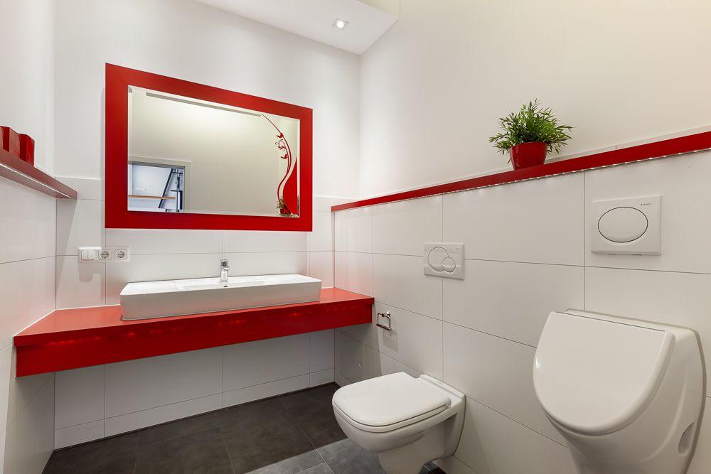 Modernes Bad mit rotem Spiegelrahmen, Waschbecken, WC, Urinal und Pflanze auf rotem Regal.