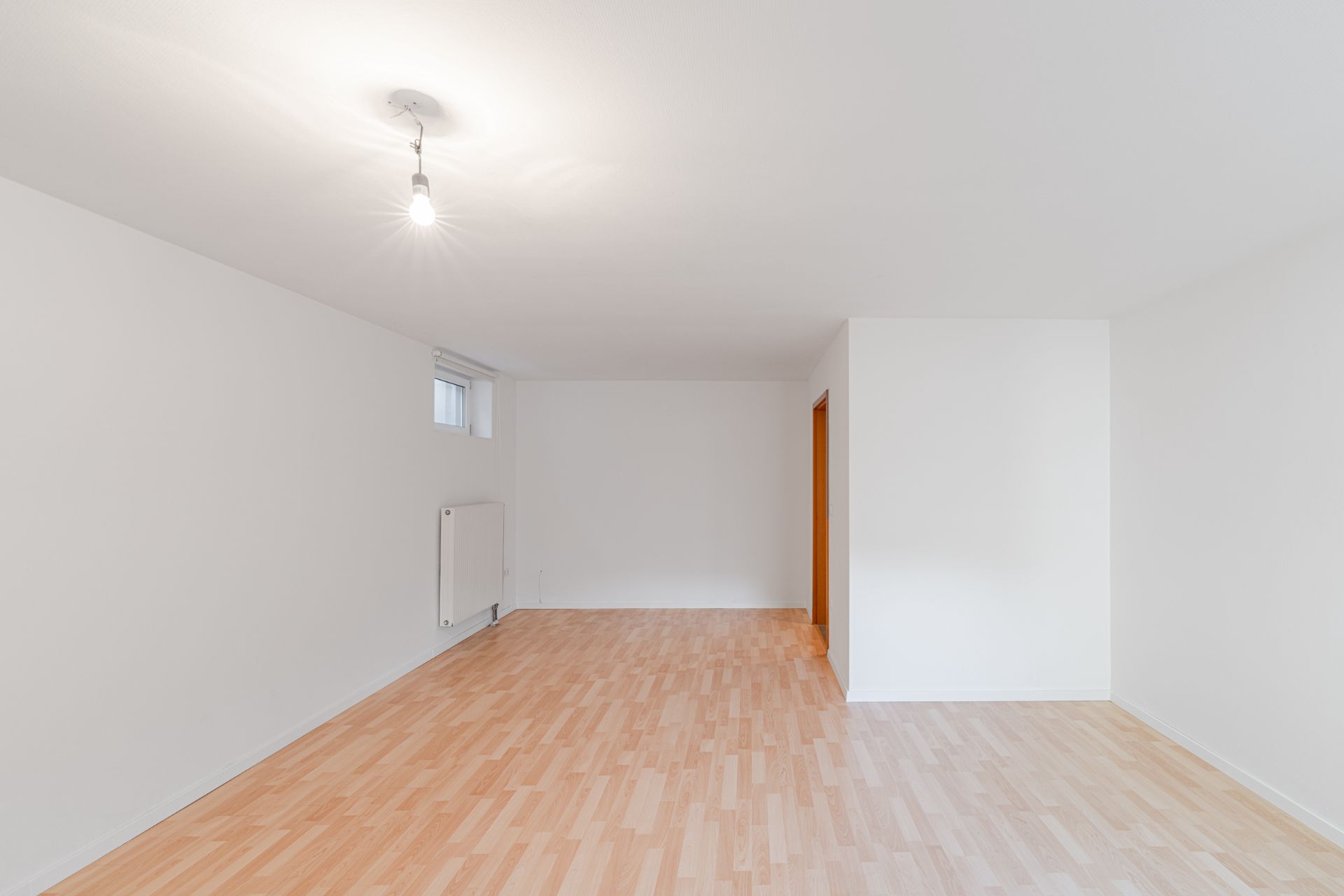 Heller, leerer Raum mit Holzfußboden, weißen Wänden, Heizung und einem kleinen Fenster.