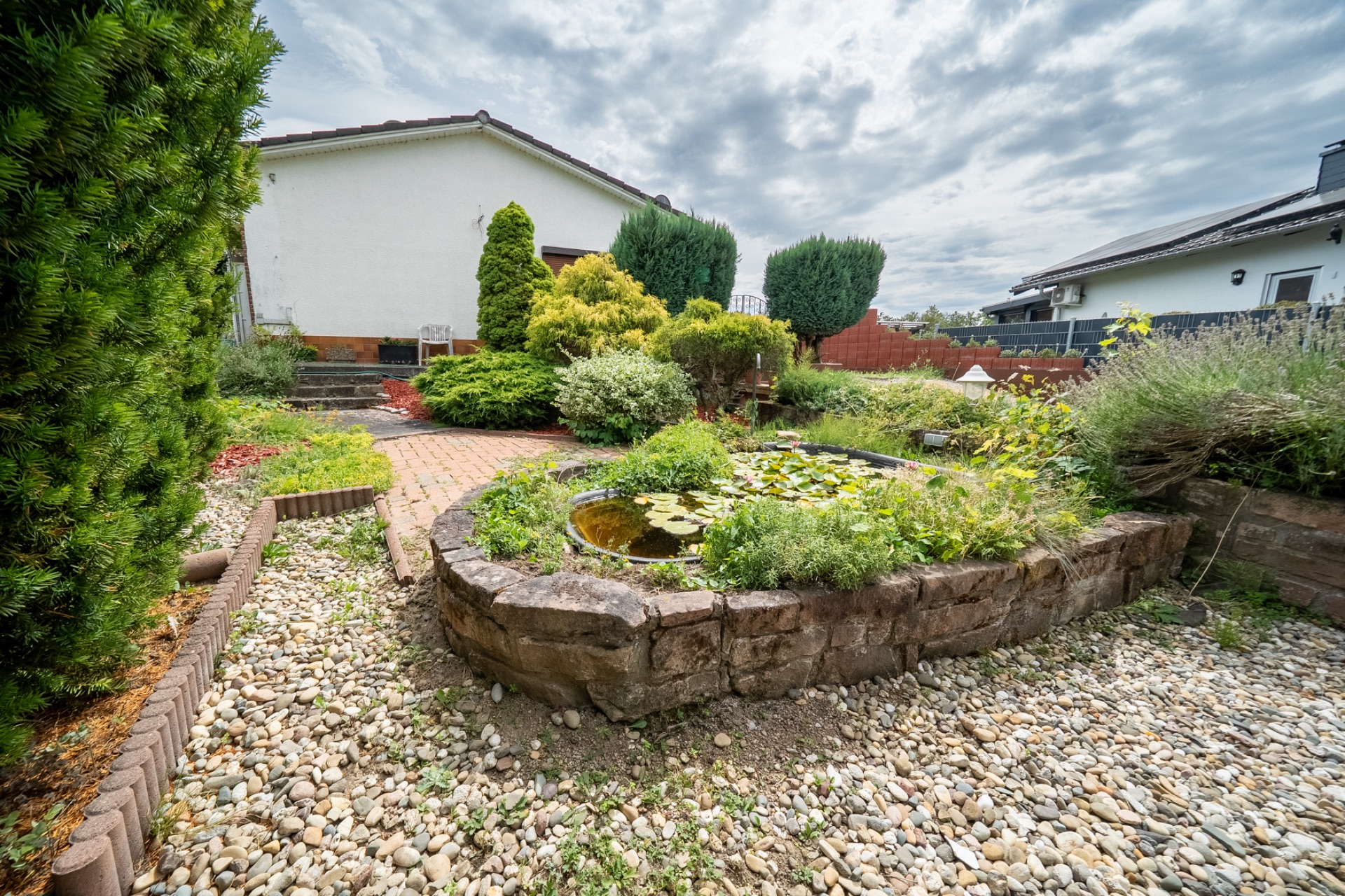 Garten mit kleinem Teich, Steinmauer und verschiedenen Büschen neben einem Wohnhaus.
