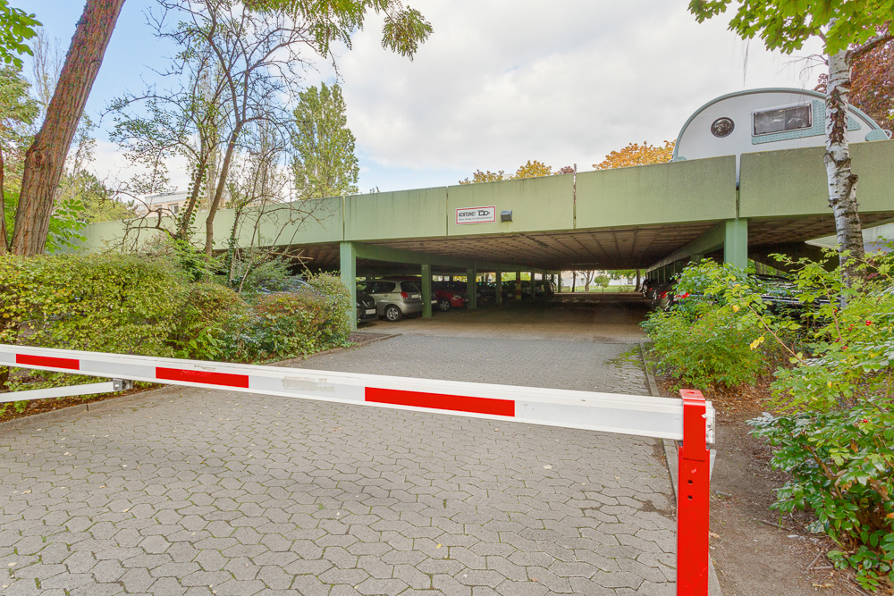 Einfahrt zu einem überdachten Parkplatz mit Schranke und umgebenden Bäumen.