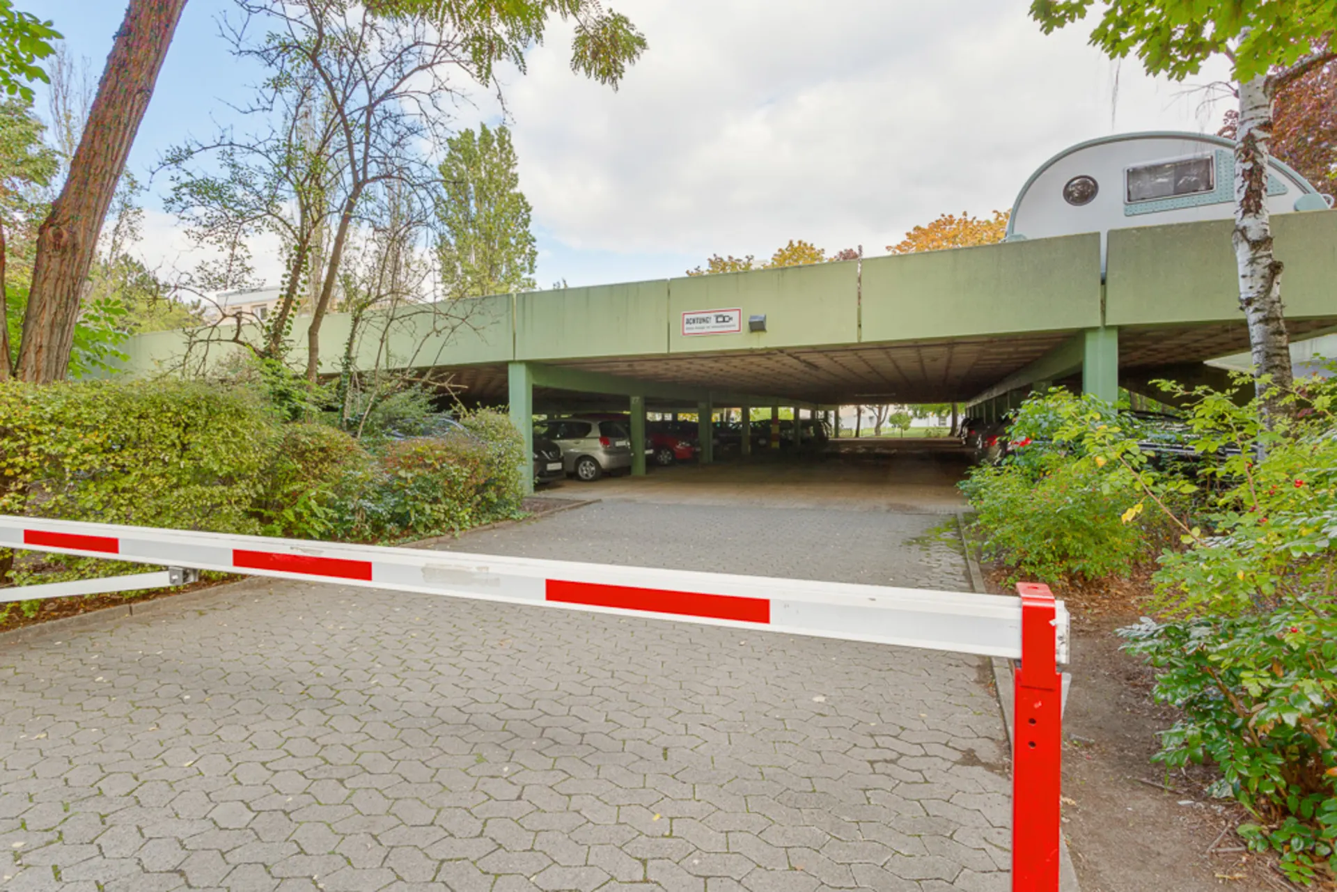Einfahrt zu einem überdachten Parkplatz mit Schranke und umgebenden Bäumen.
