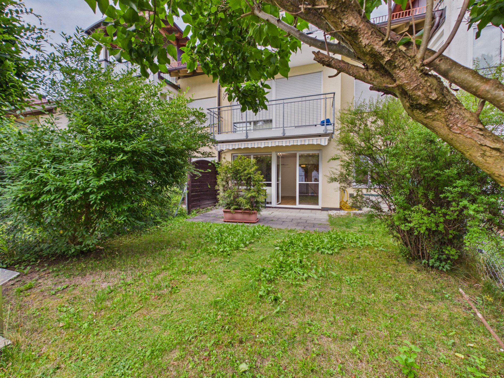 Garten mit Rasen, Sträuchern und Terrasse vor einem Wohnhaus mit großen Fenstern.