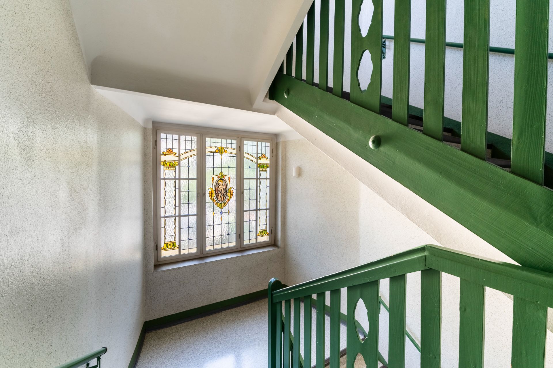 Treppenhaus mit grünem Geländer und buntem Bleiglasfenster.