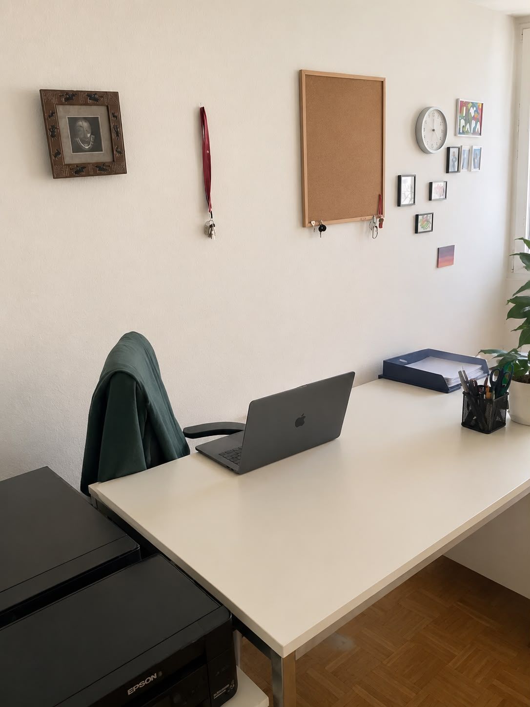 Schreibtisch mit Laptop und Pflanze, Wand mit Uhr, Pinnwand und Bildern.