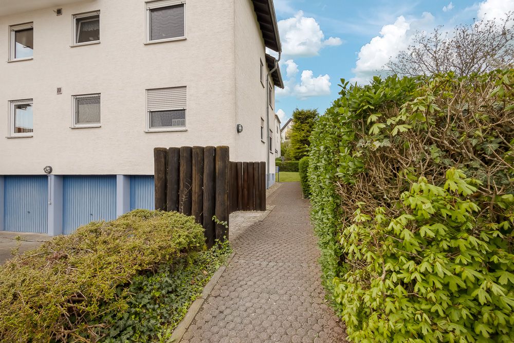 Gepflasterter Weg neben einem Wohnhaus mit blauen Garagentoren und grüner Hecke.