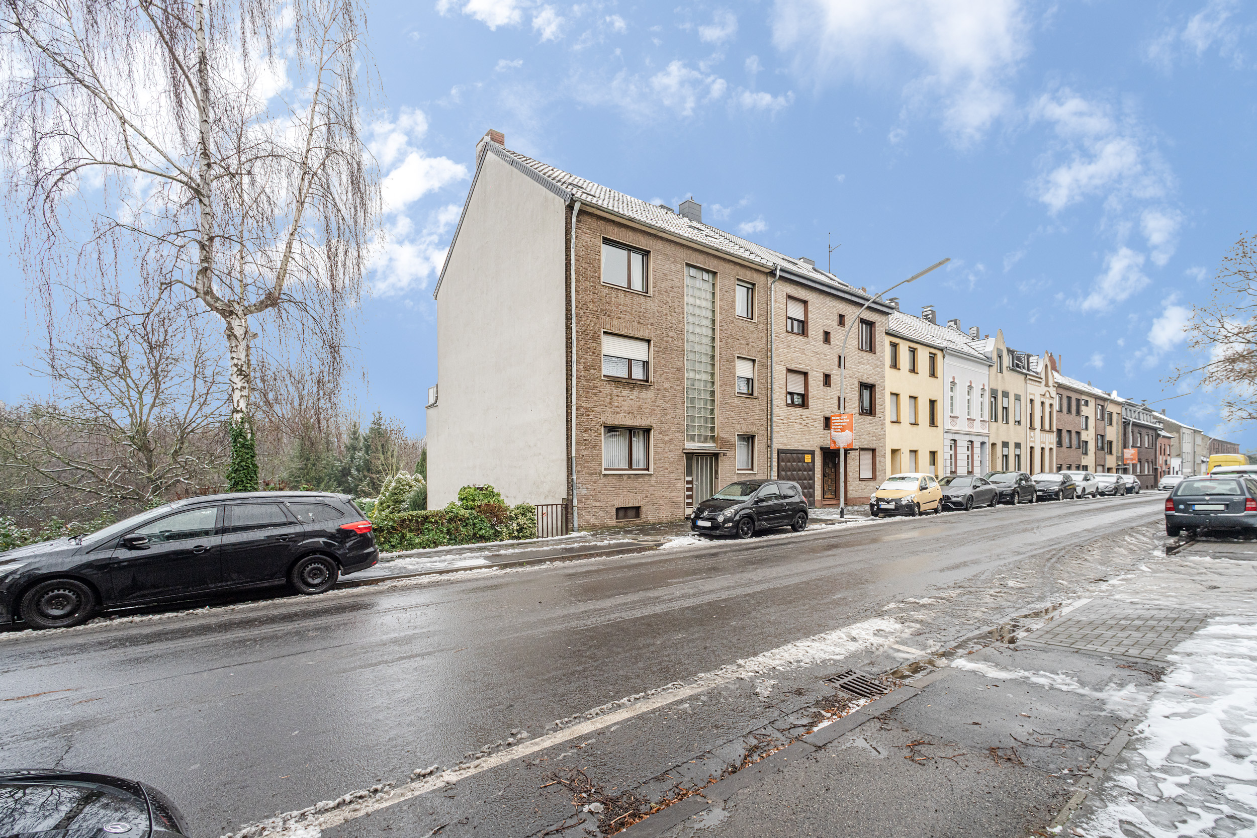 Mehrfamilienhaus an einer verschneiten Straße mit parkenden Autos bei Tageslicht.