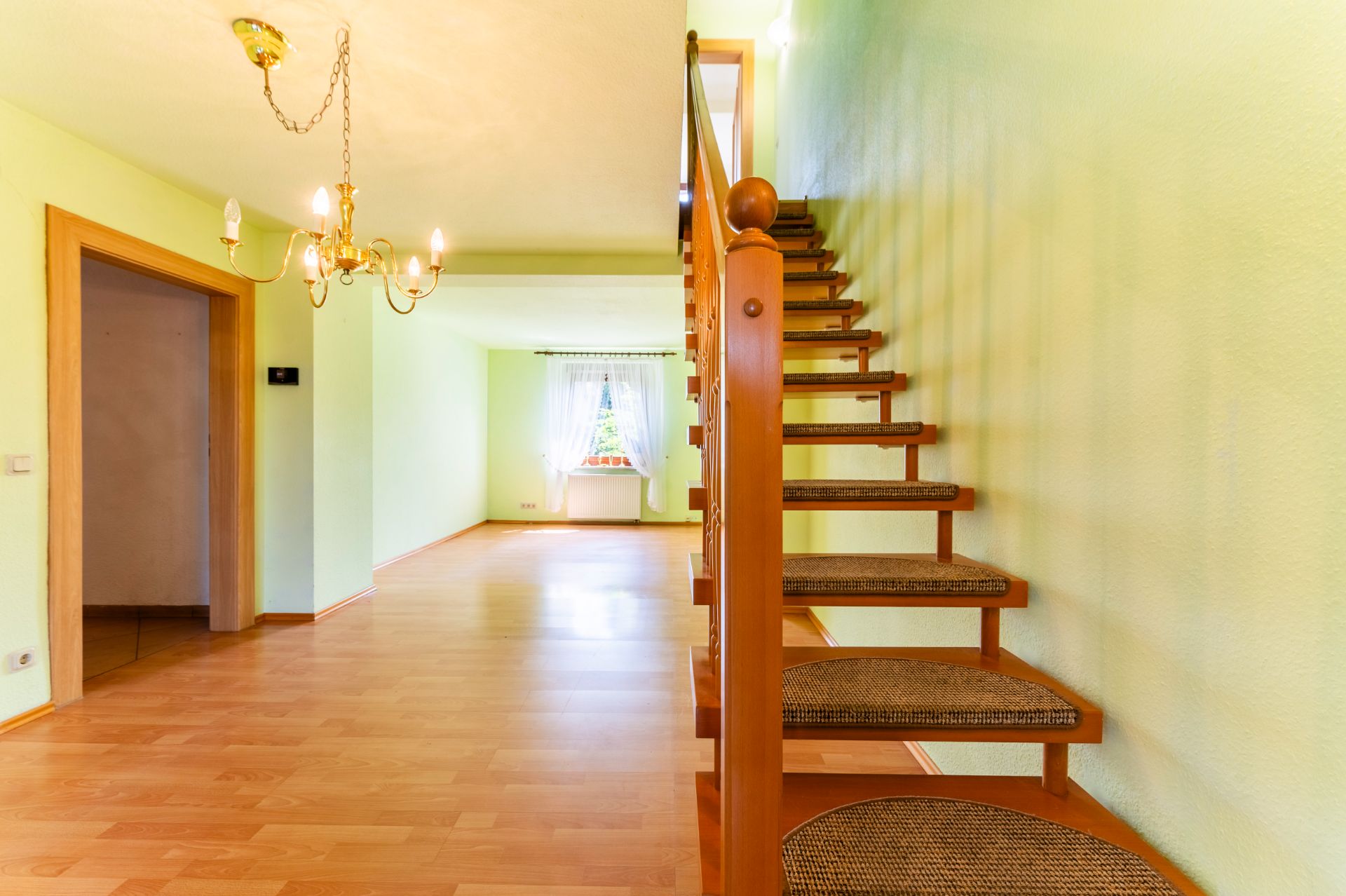 Helles Wohnzimmer mit Holztreppe, Kronleuchter und großem Fenster mit weißen Vorhängen.