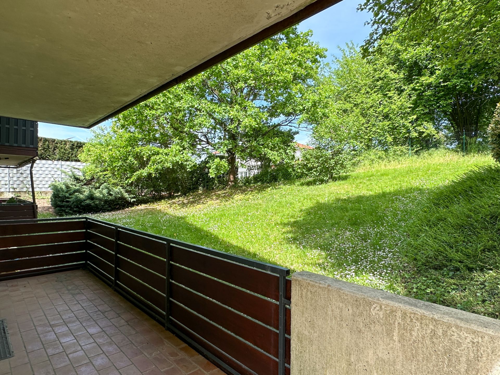 Überdachte Terrasse mit braunem Geländer, Blick auf grünen Garten mit Bäumen und Rasen.