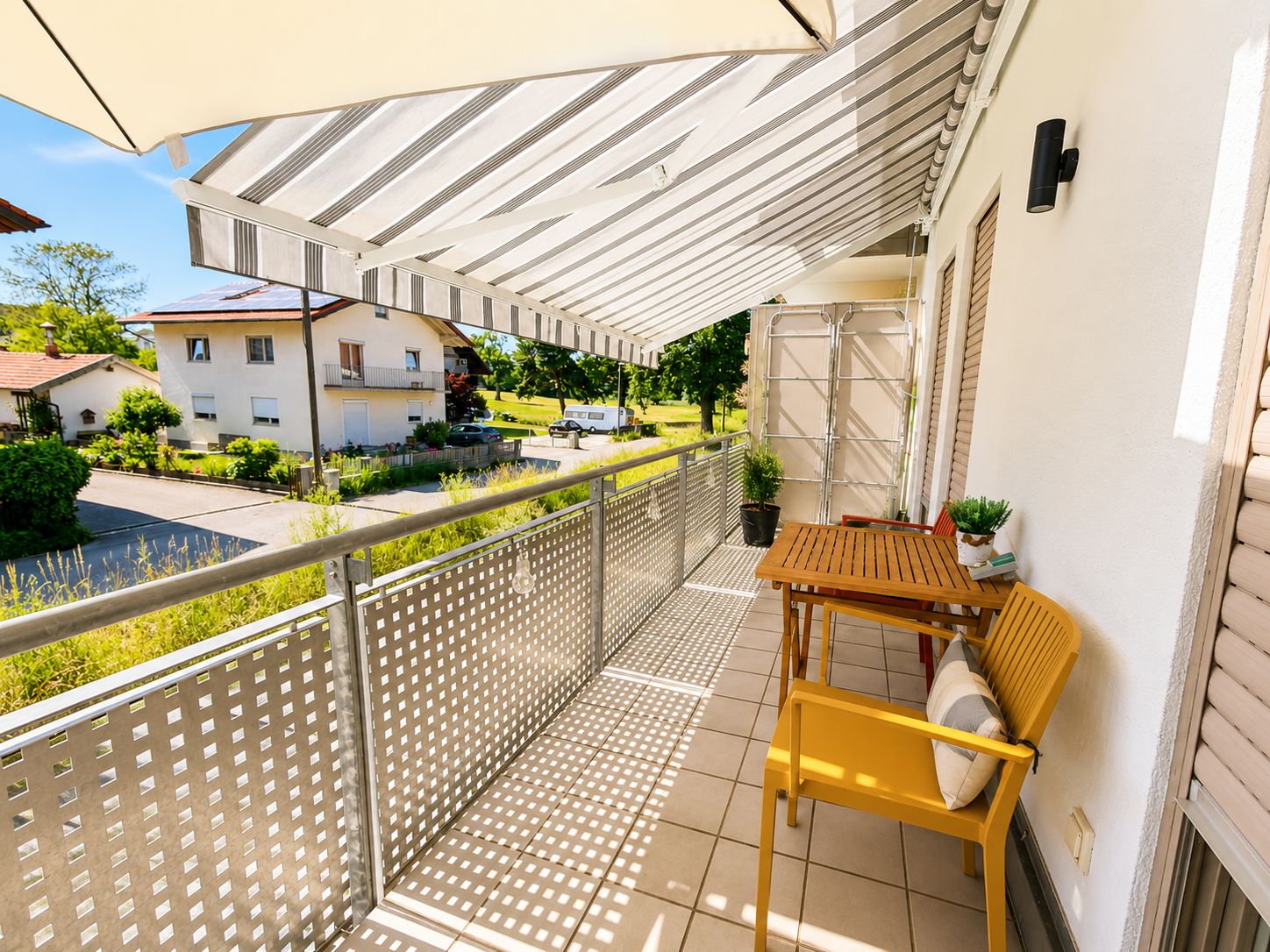 Balkon mit Markise, Tisch, Stuhl und Pflanzen, Blick auf Nachbarhäuser und Straße.