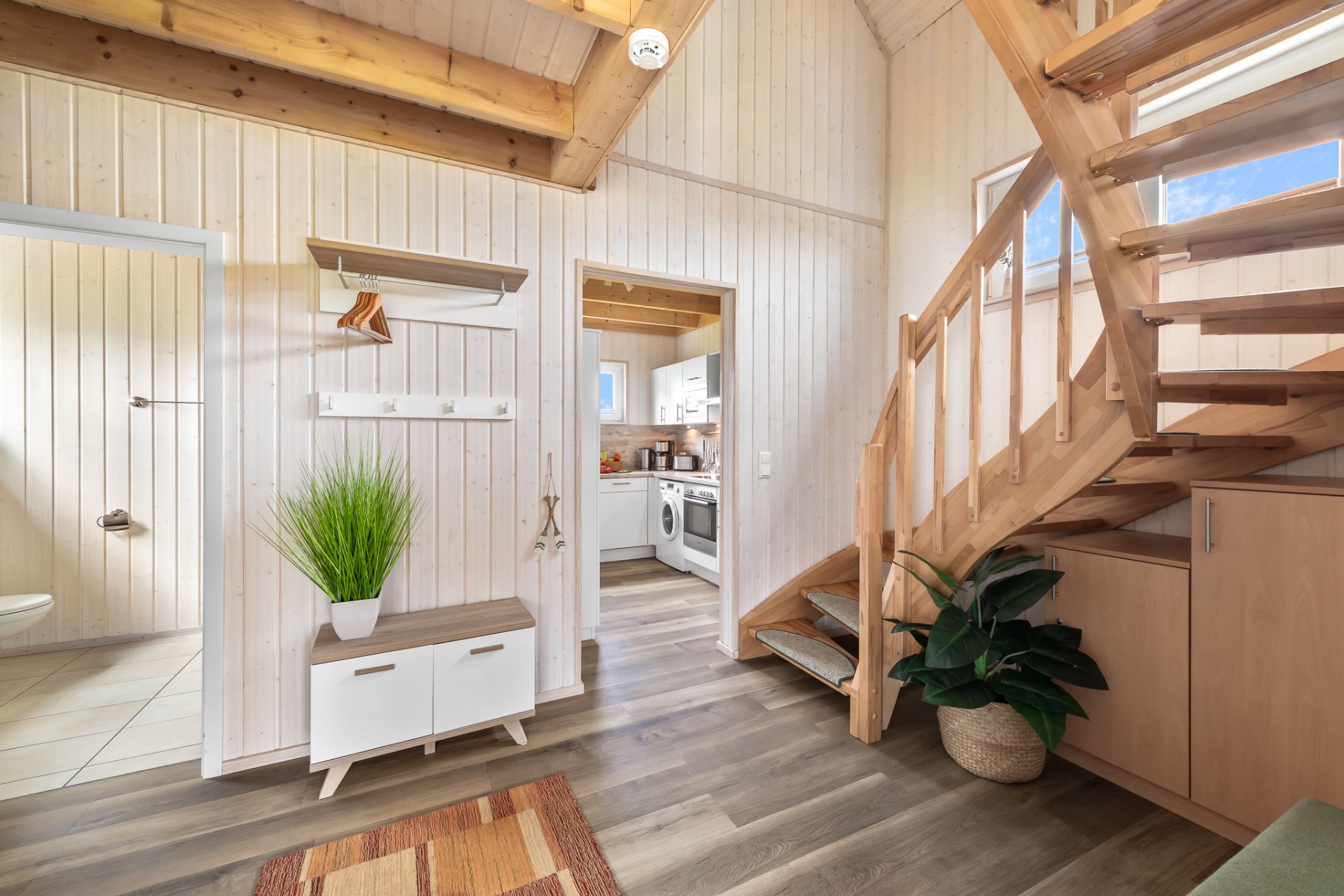 Heller Flur mit Holztreppe, Blick ins Bad und in eine helle Küche, Pflanze und Garderobe sichtbar.