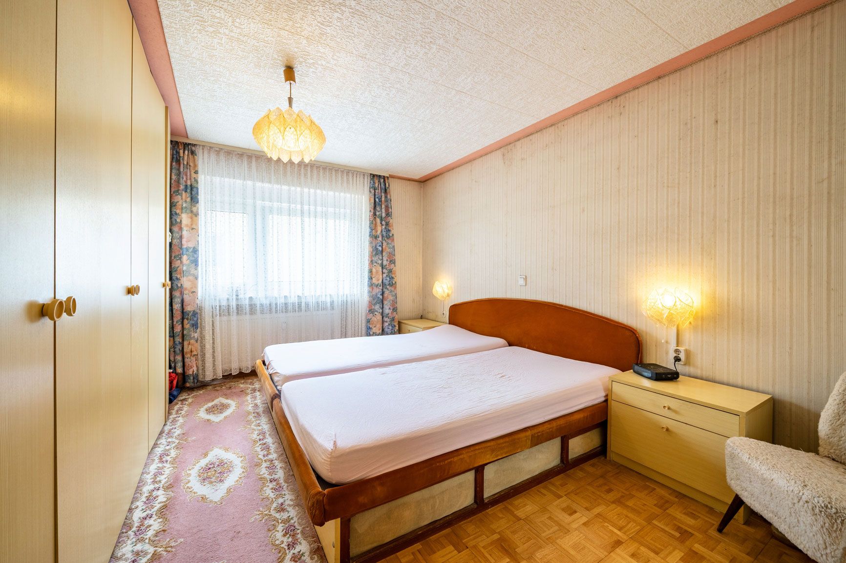 Schlafzimmer mit zwei Betten, Nachttischen, Kleiderschrank und großem Fenster mit Vorhängen.