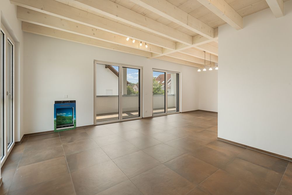 Helles, leeres Zimmer mit großen Fenstern, braunen Fliesen und einem kleinen Aquarium an der Wand.
