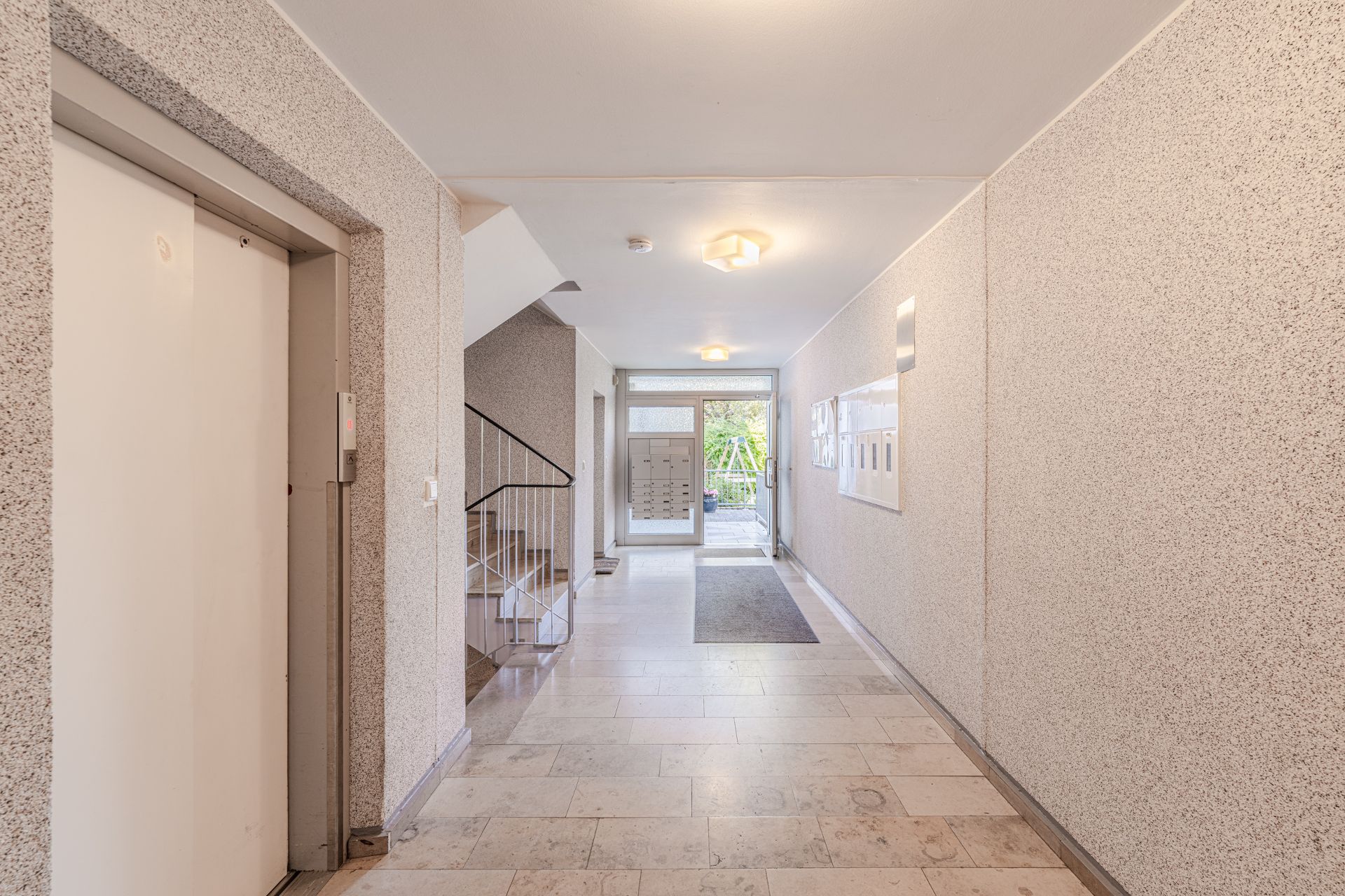 Heller Wohnungseingang mit Aufzug, Treppe, Briefkästen und Glastür zum Außenbereich.