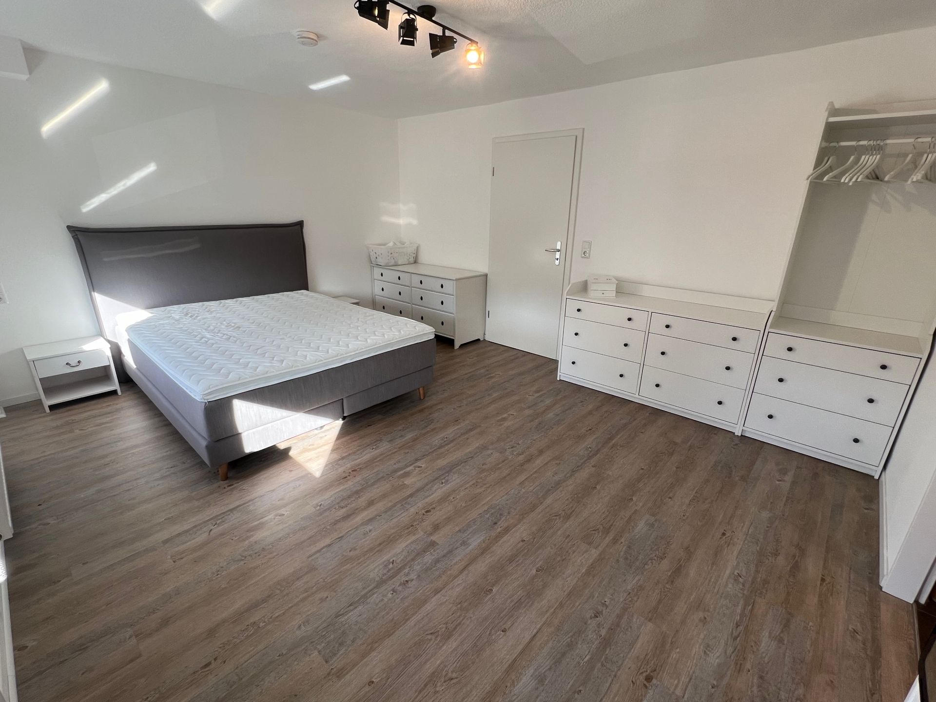 Helles Schlafzimmer mit Doppelbett, Nachttisch, Kommoden und offenem Kleiderschrank auf Holzfußboden.