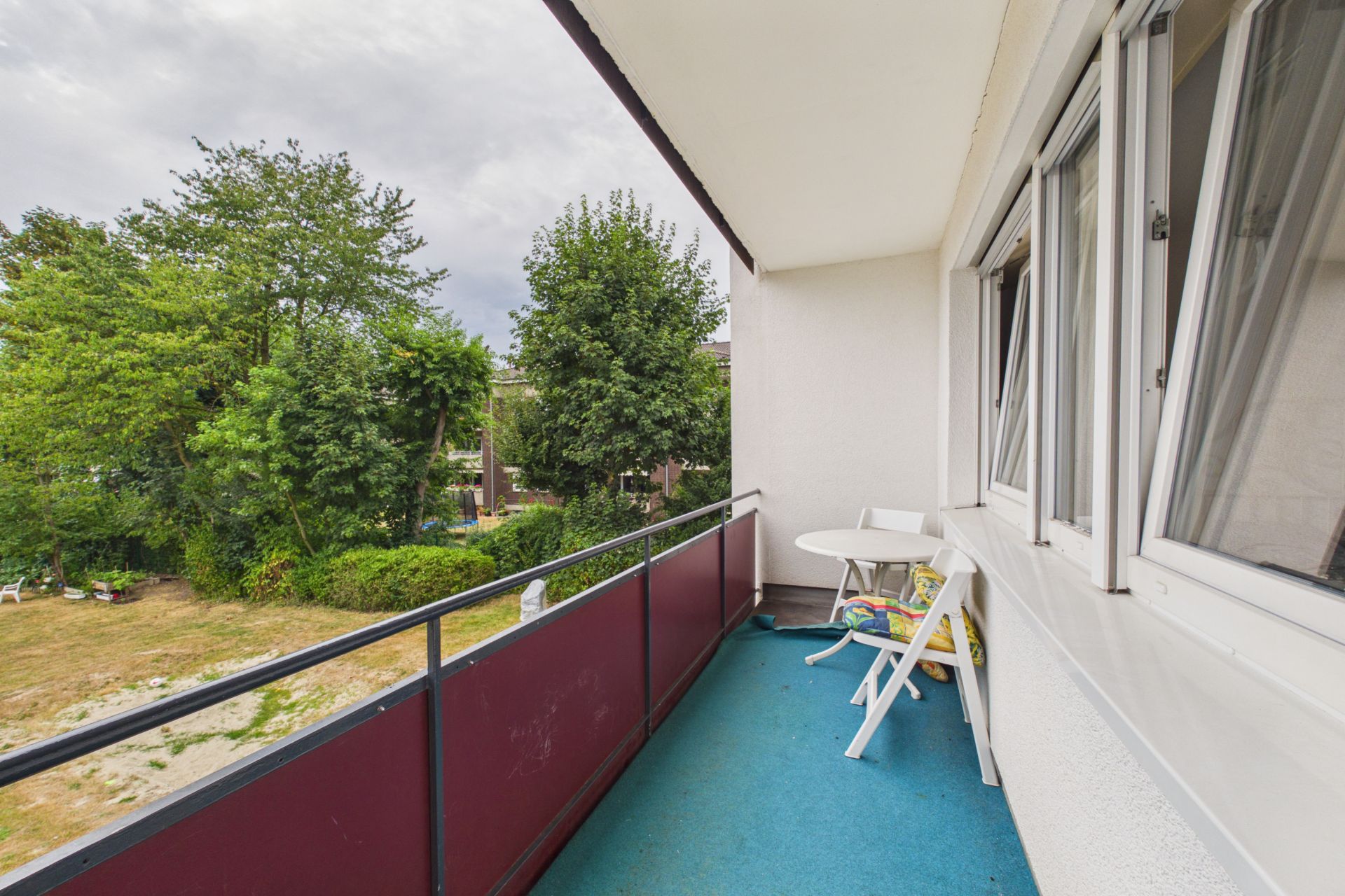 Balkon mit blauem Boden, weißem Tisch und zwei Stühlen, Blick auf grüne Bäume.