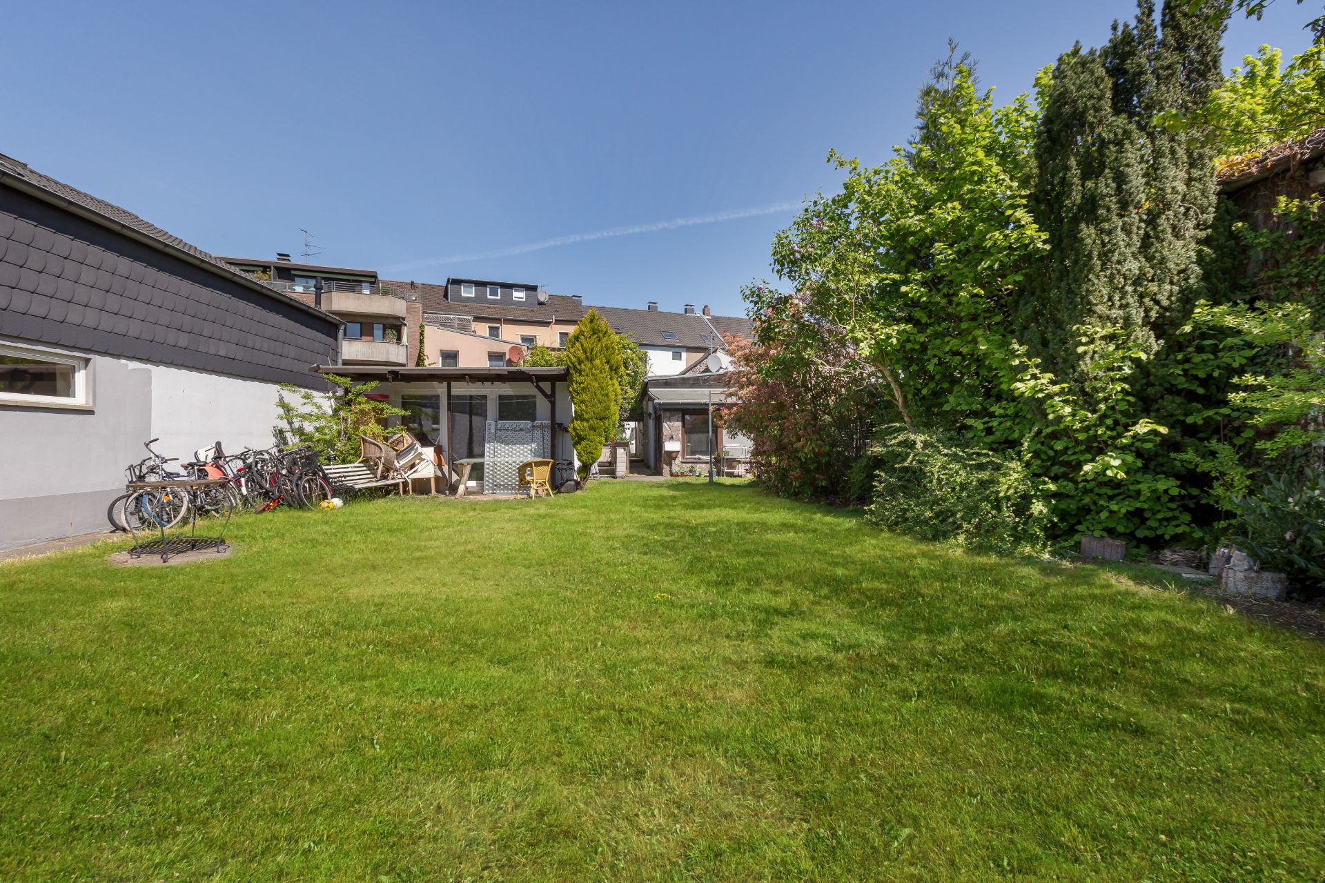 Grüner Garten mit Rasen, Bäumen, Fahrrädern und Gartenmöbeln hinter einem Wohnhaus.