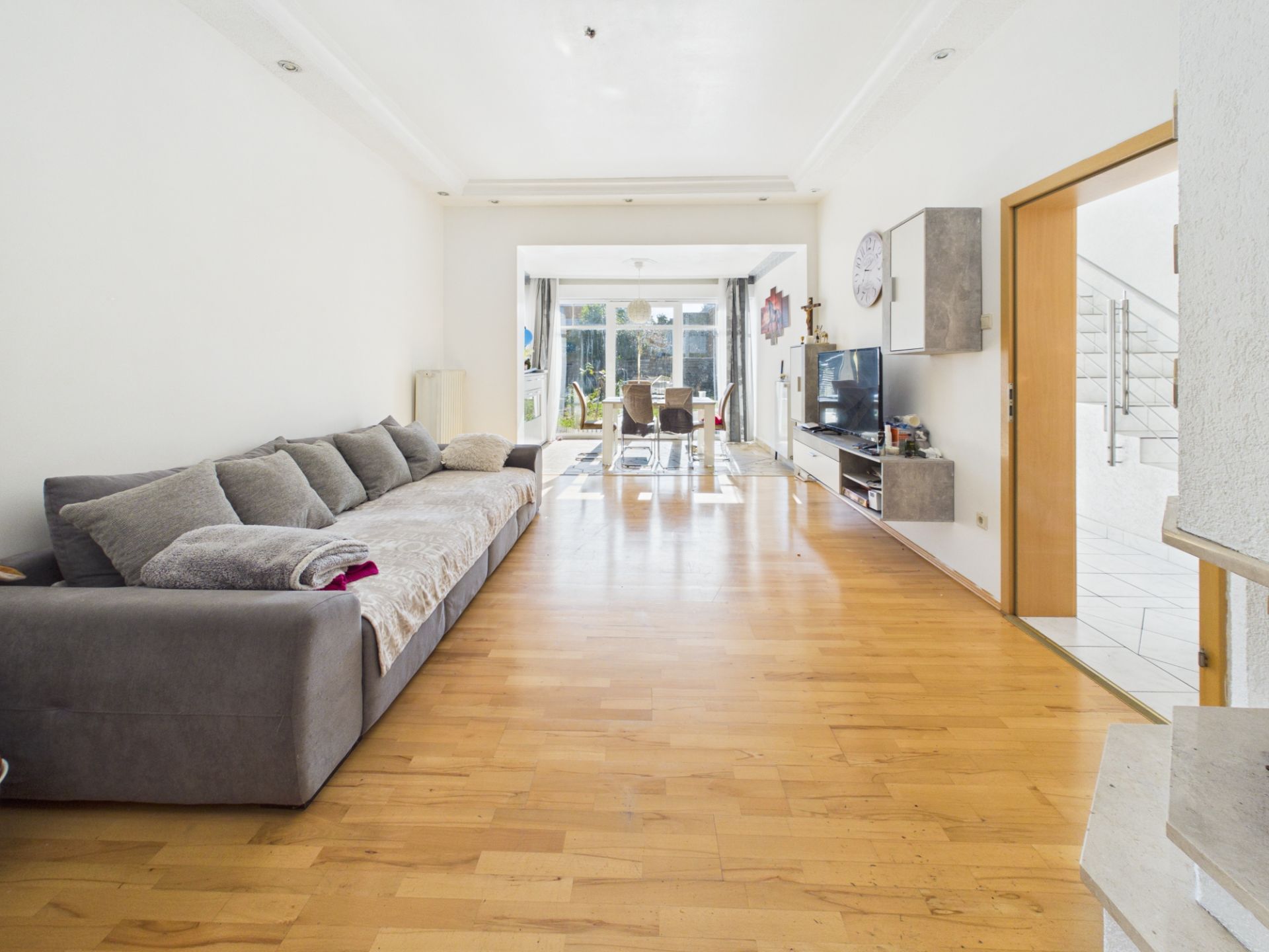 Wohnzimmer mit grauem Sofa, Holzboden, Fernseher und Blick auf Essbereich mit Fenstern.