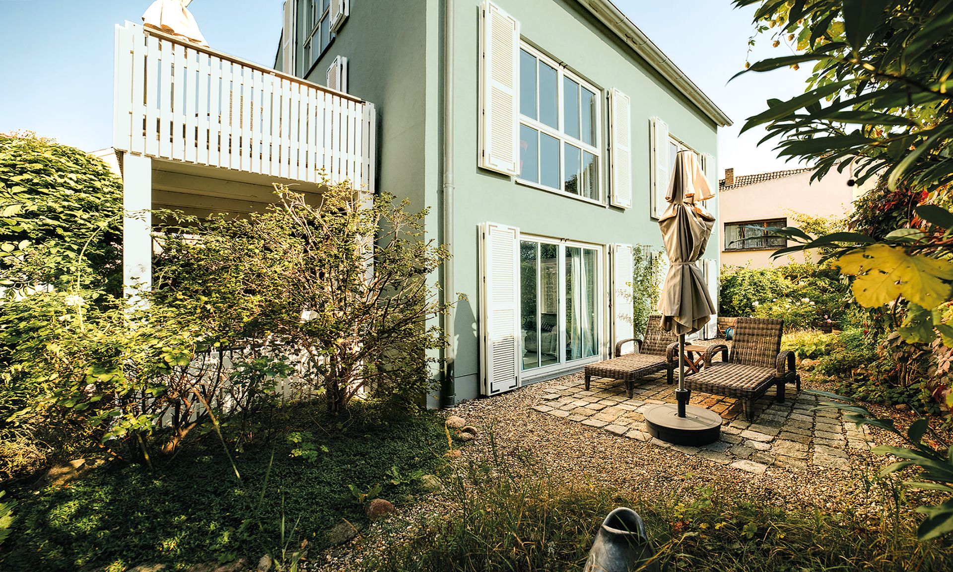 Grünes Wohnhaus mit Terrasse, Gartenmöbeln und vielen Pflanzen im Garten.