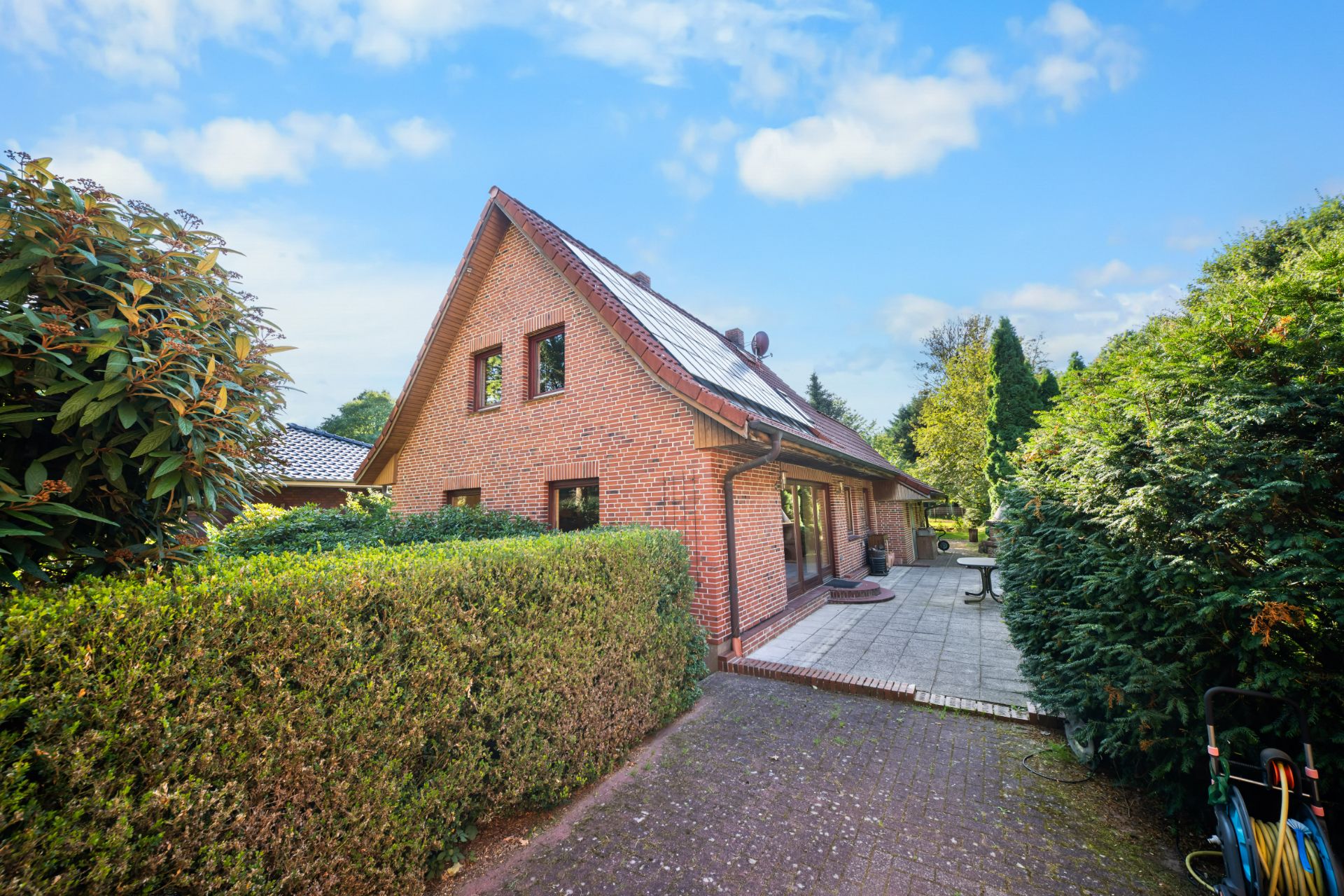 Backsteinhaus mit Satteldach, Terrasse und von Hecken eingefasstem Gartenweg.