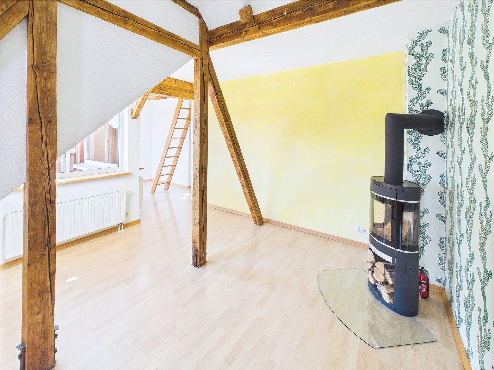 Heller Raum mit Holzofen, Holzbalken, gelber Wand und Tapete mit Blättermuster.