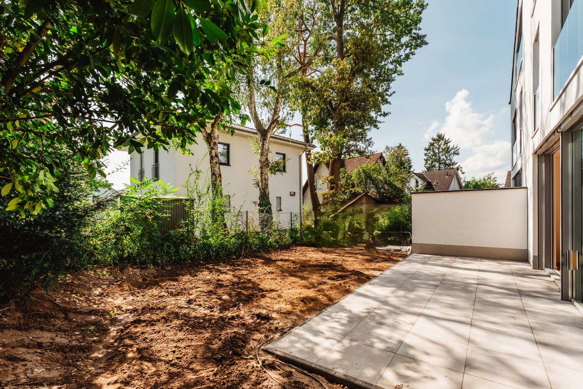 Gepflasterte Terrasse neben einem unbepflanzten Garten, umgeben von Bäumen und Wohnhäusern.