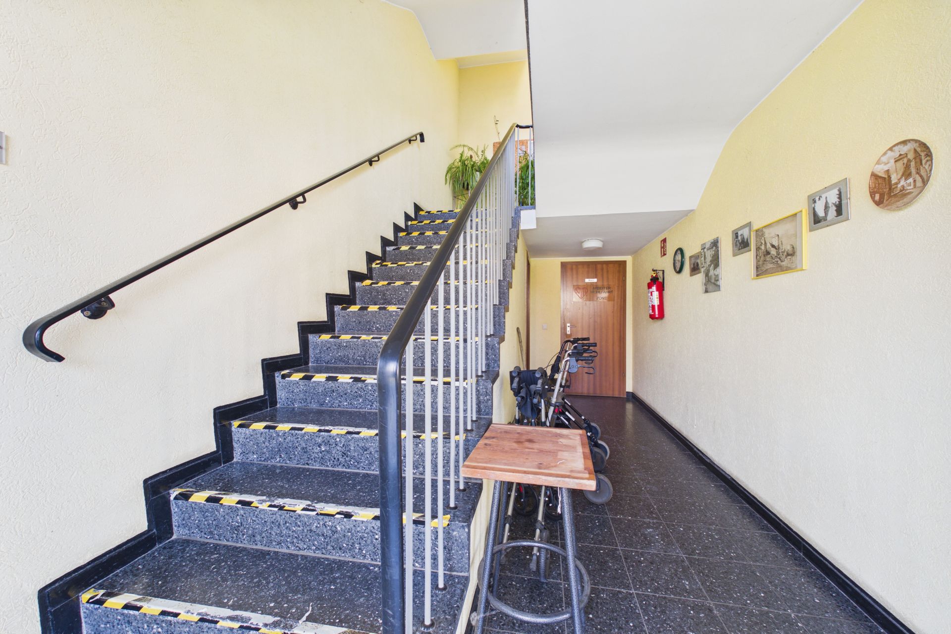 Hausflur mit Treppe, Bildern an der Wand und einigen Rollatoren neben einem Tisch.