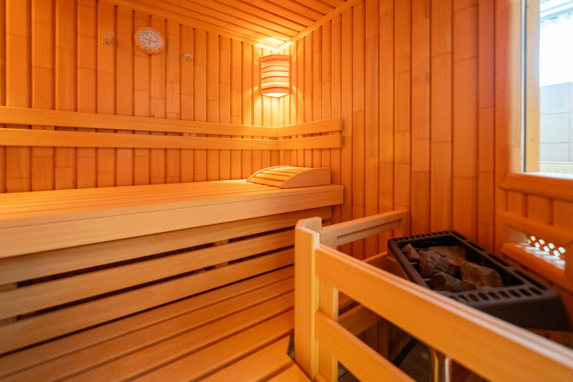 Holzsauna mit Bank, Kopfstütze, Ofen und Wanduhr, beleuchtet von einer Lampe.