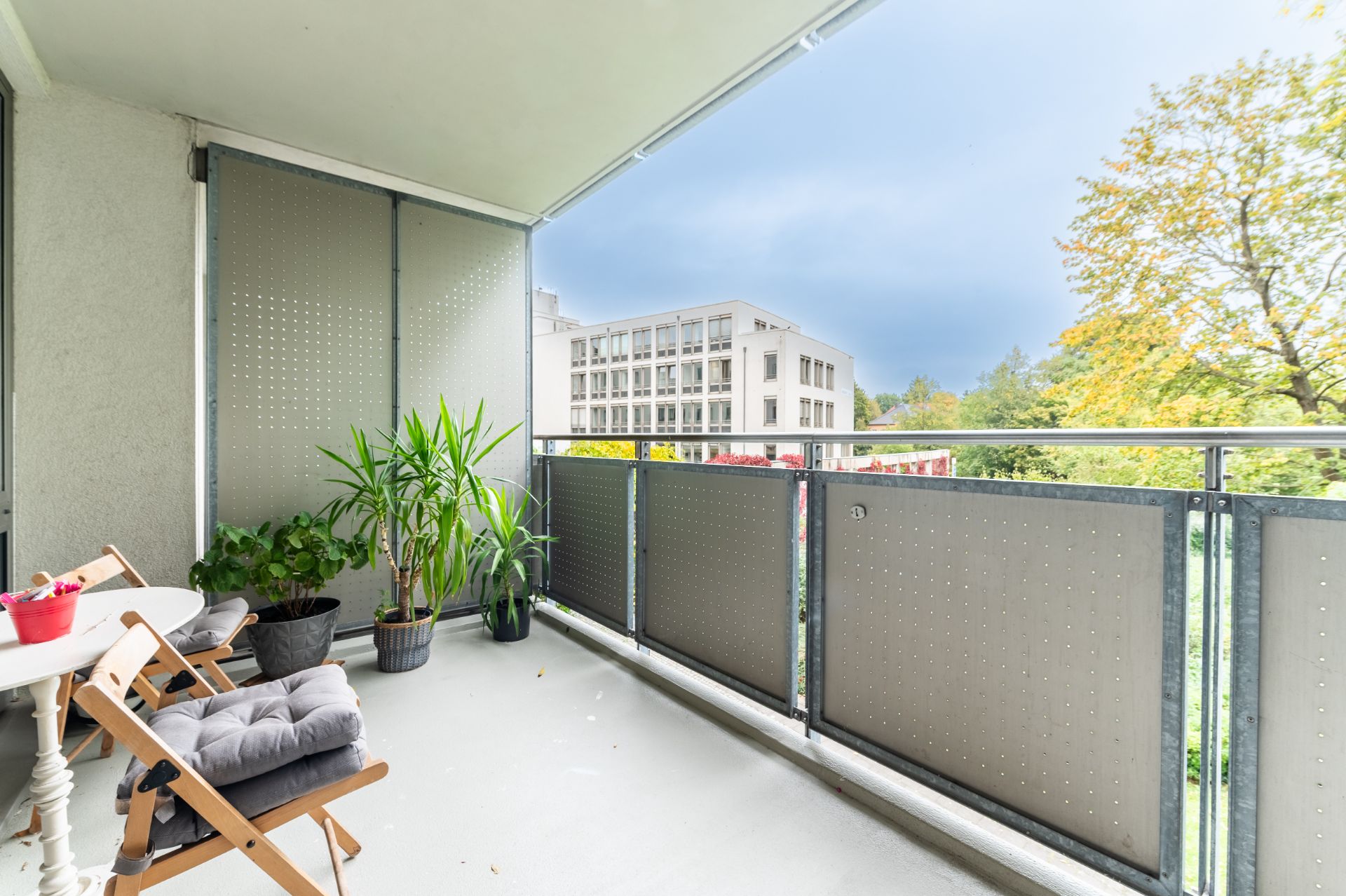Balkon mit Stühlen, Tisch, Topfpflanzen und Blick auf Bäume sowie ein Gebäude im Hintergrund.