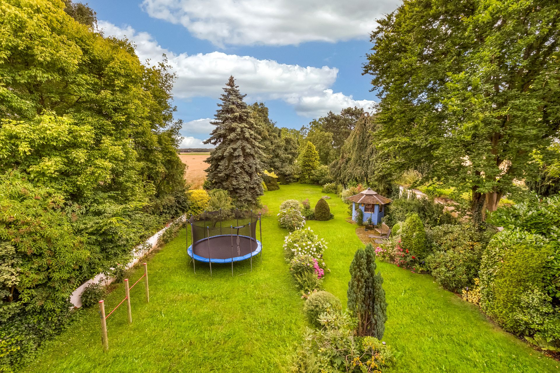 Großer grüner Garten mit Trampolin und blauem Pavillon, umgeben von Bäumen und Sträuchern.