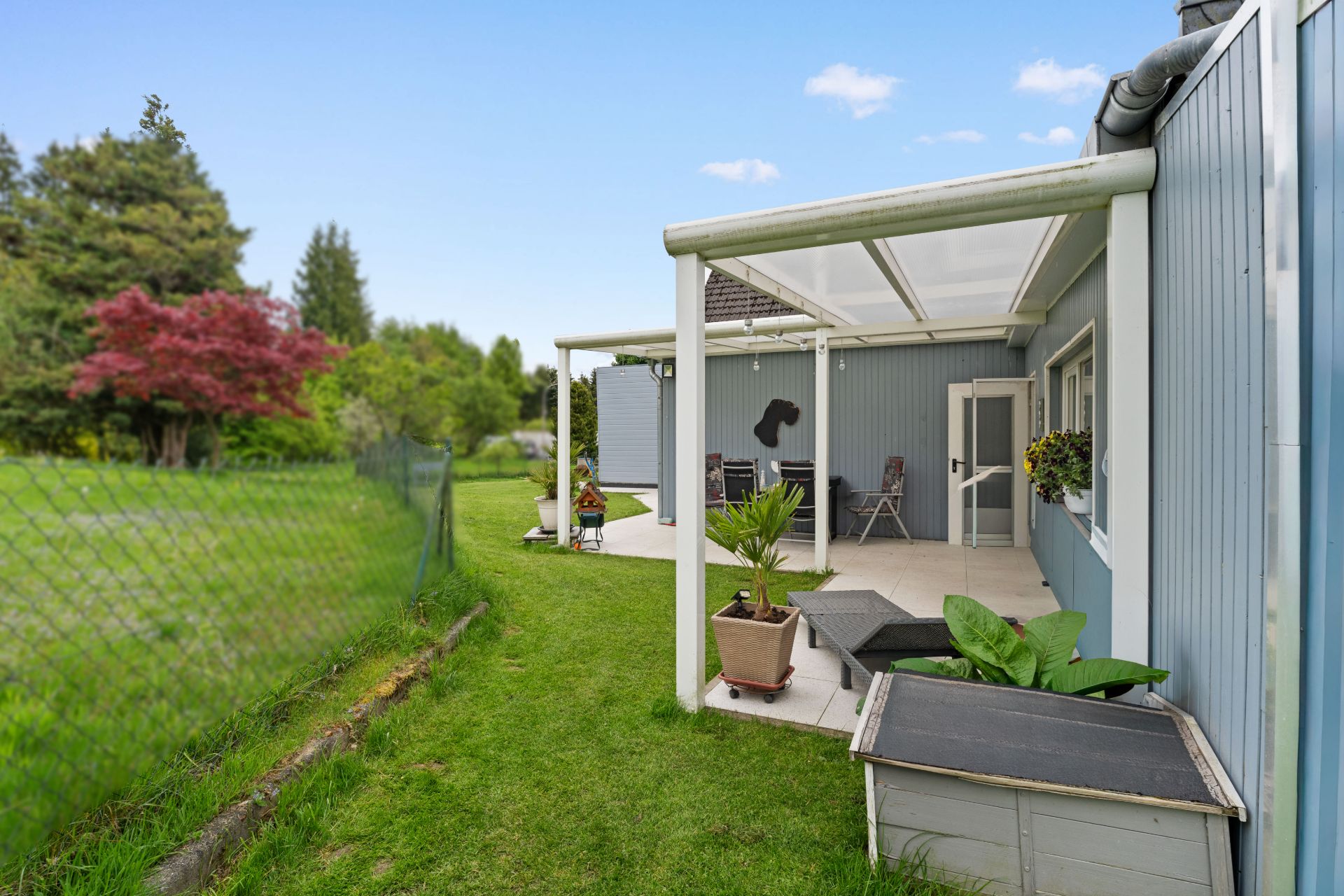 Überdachte Terrasse mit Garten, Pflanzenkübeln und angrenzender Rasenfläche neben einem Haus.