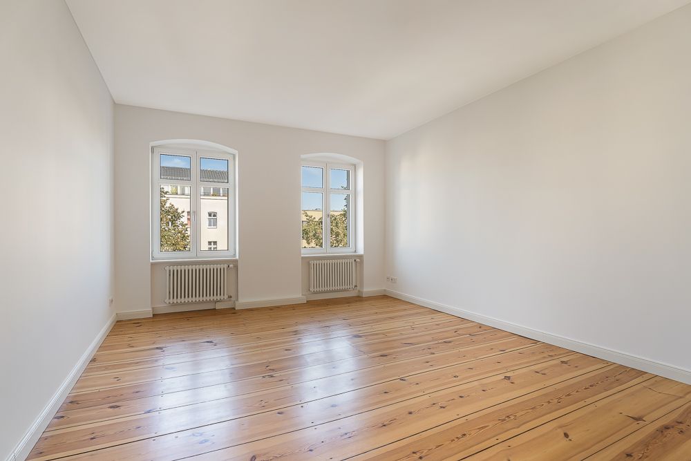 Heller leerer Raum mit Holzfußboden und zwei Fenstern an der Stirnwand.