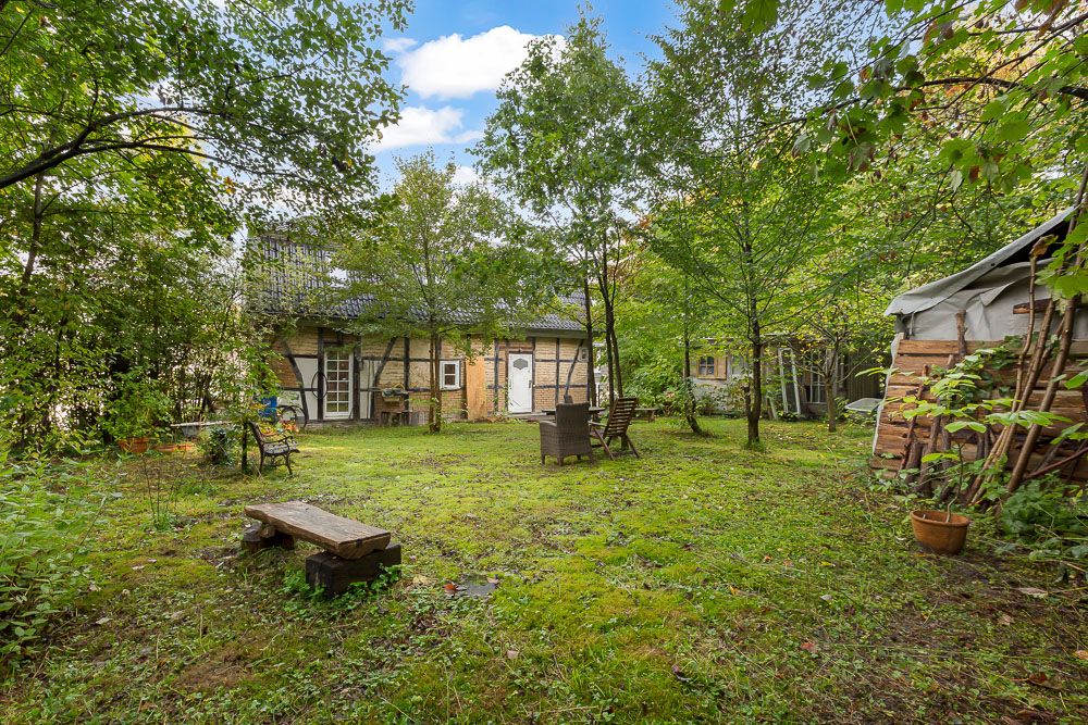Grüner Garten mit Bäumen, Sitzbänken und einem Fachwerkhaus im Hintergrund.