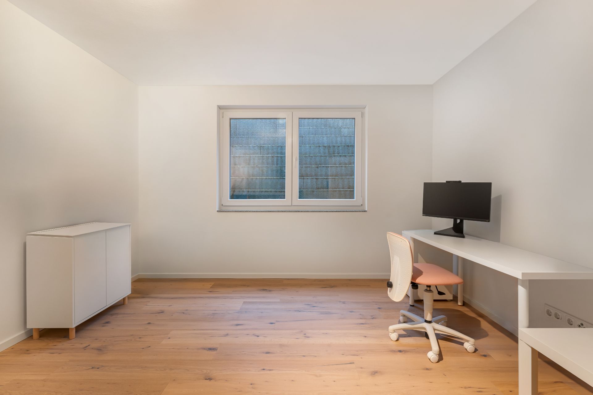Helles Zimmer mit Fenster, weißem Schreibtisch, Stuhl, Monitor und weißem Sideboard auf Holzboden.